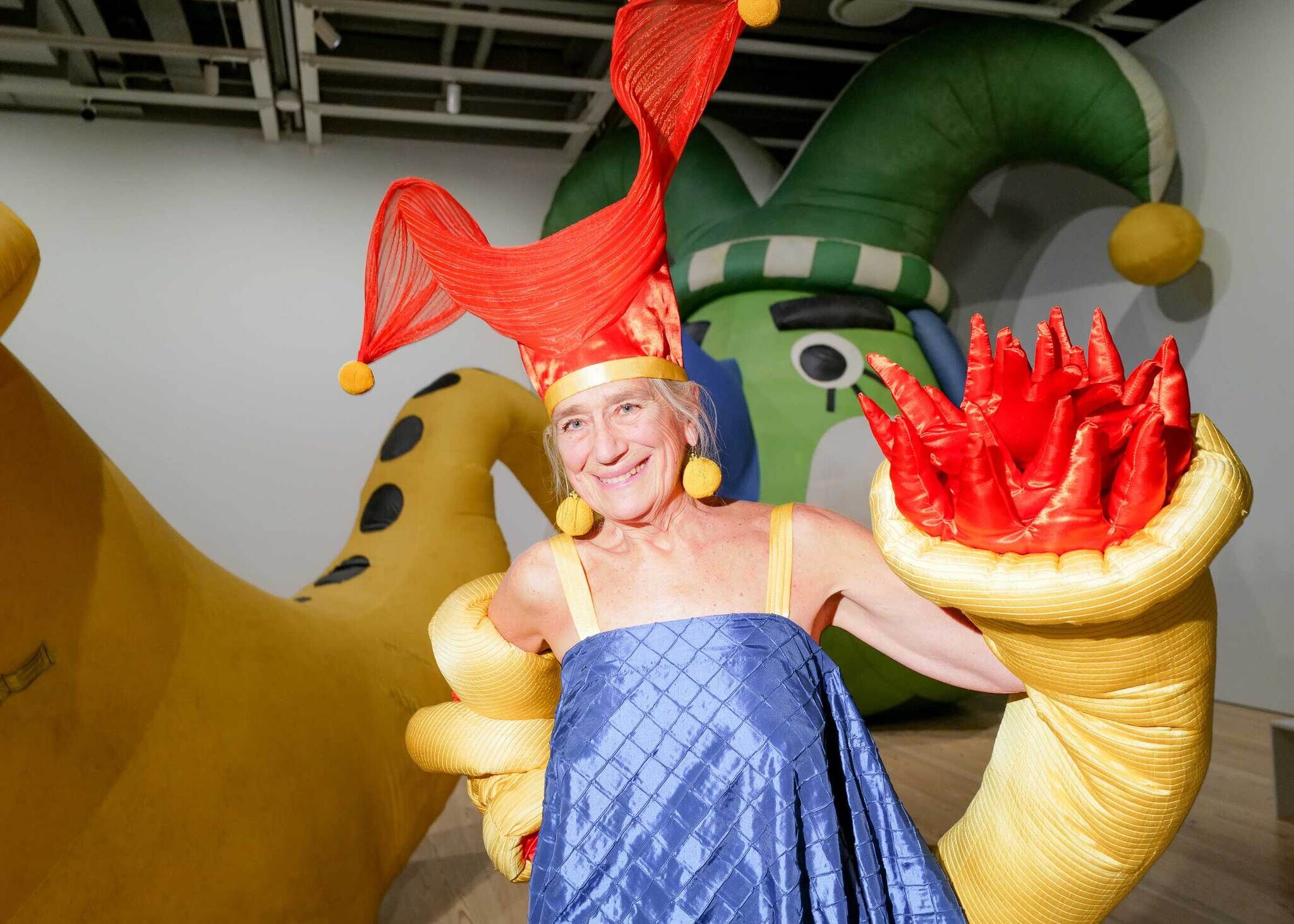 Artist Pat Oleszko stands in a gallery wearing a bright red jester-style hat and sculptural costume with oversized yellow sleeves with protruding red forms. Behind her is her large inflatable sculpture depicting a green figure blowing a yellow trumpet, installed in the gallery space with wood floors and white walls