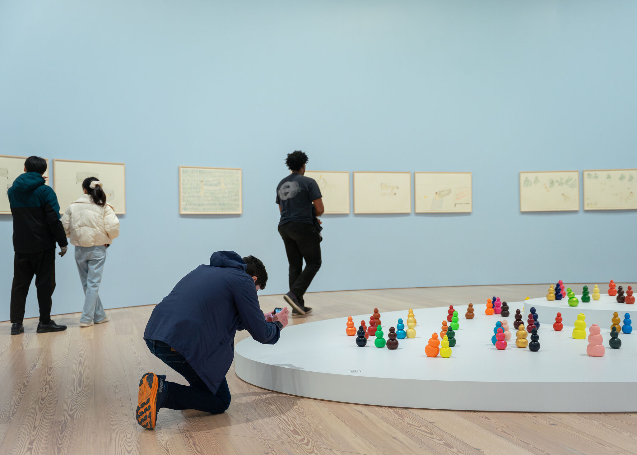 A person kneels to photograph colorful small sculptures displayed on a low white platform in a gallery.