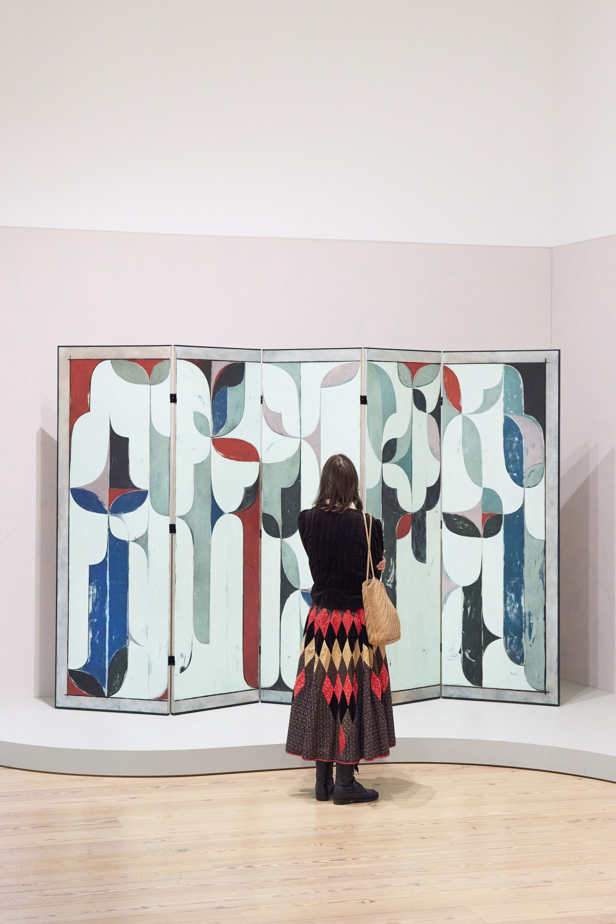 A woman stands facing a large colorful abstract folding screen in a gallery.