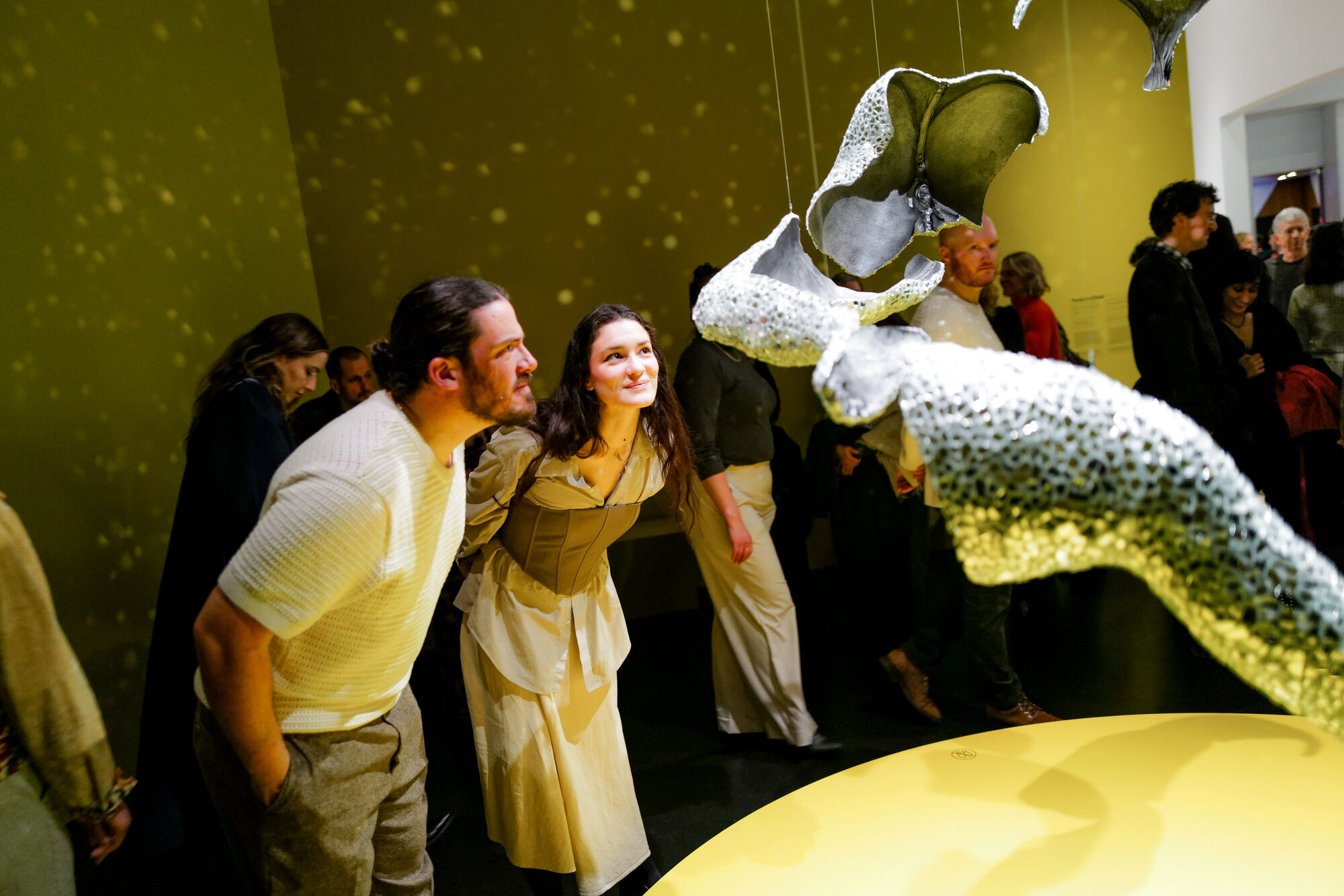 A man and woman lean forward to closely examine suspended porous sculptures at a gallery.