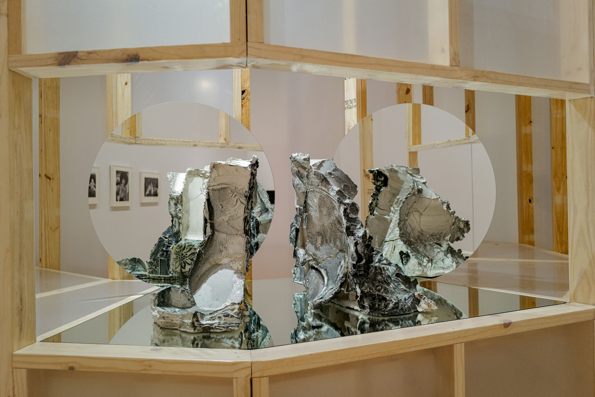 Two broken metallic sculptures sit on a mirrored shelf framed by circular openings and wooden supports.