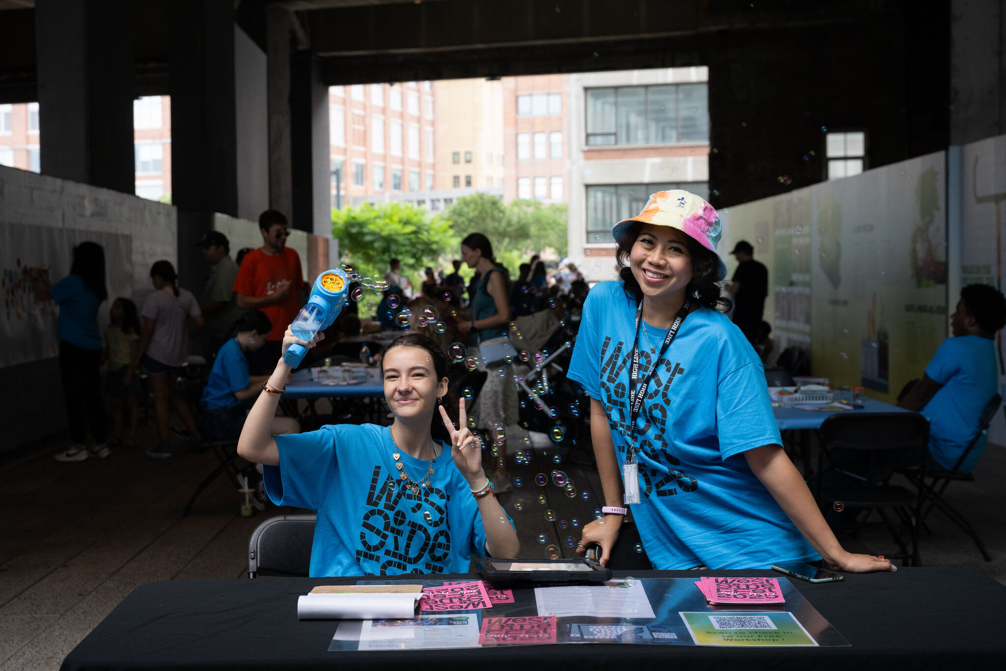 Two smiling volunteers in blue shirts blow bubbles and greet visitors at an outdoor information table.