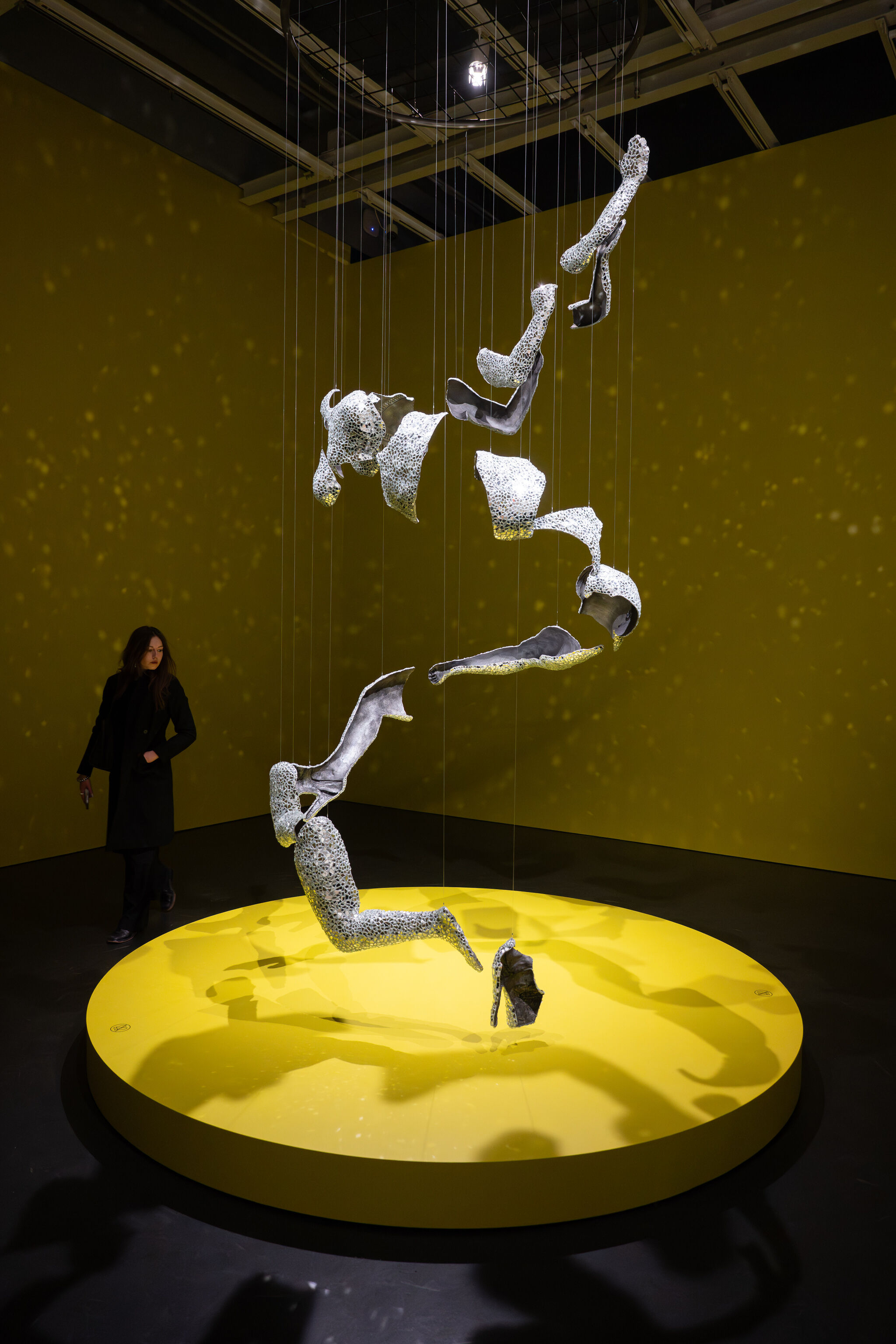Abstract perforated metal forms hang from wires above a lit yellow circular platform in a gallery.