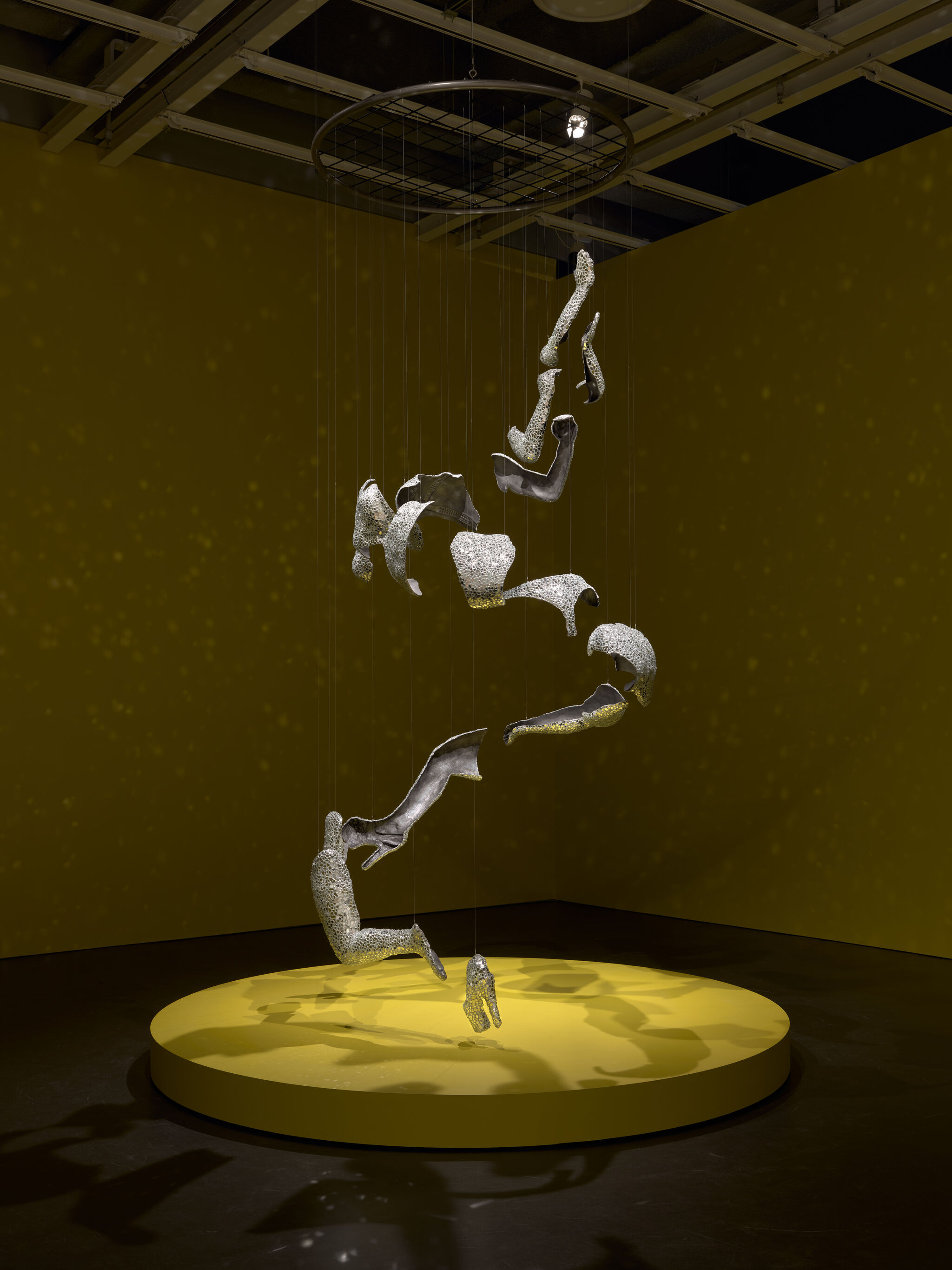 A spiral of fragmented metallic human forms hangs suspended above a round yellow platform.