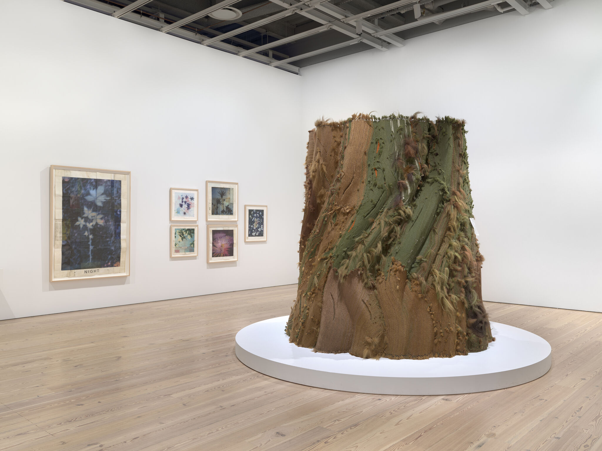 Large frayed green and brown textile sculpture resembling a tree stump displayed on a white round platform.