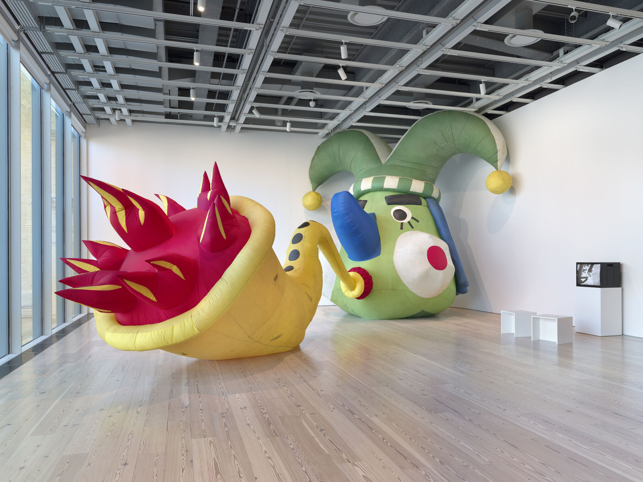 Large inflatable green jester head and connected yellow-red spiky inflatable lie across a gallery floor.