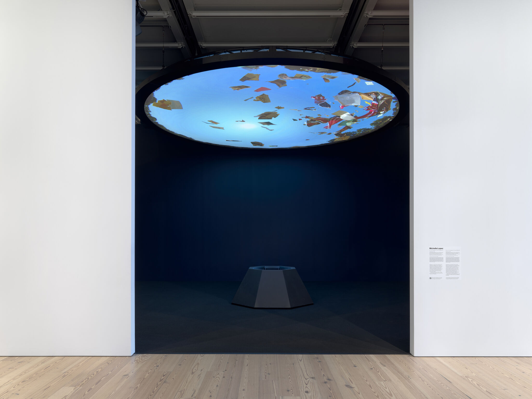 Circular ceiling screen displays floating colorful debris above a dark gallery space with central pedestal.