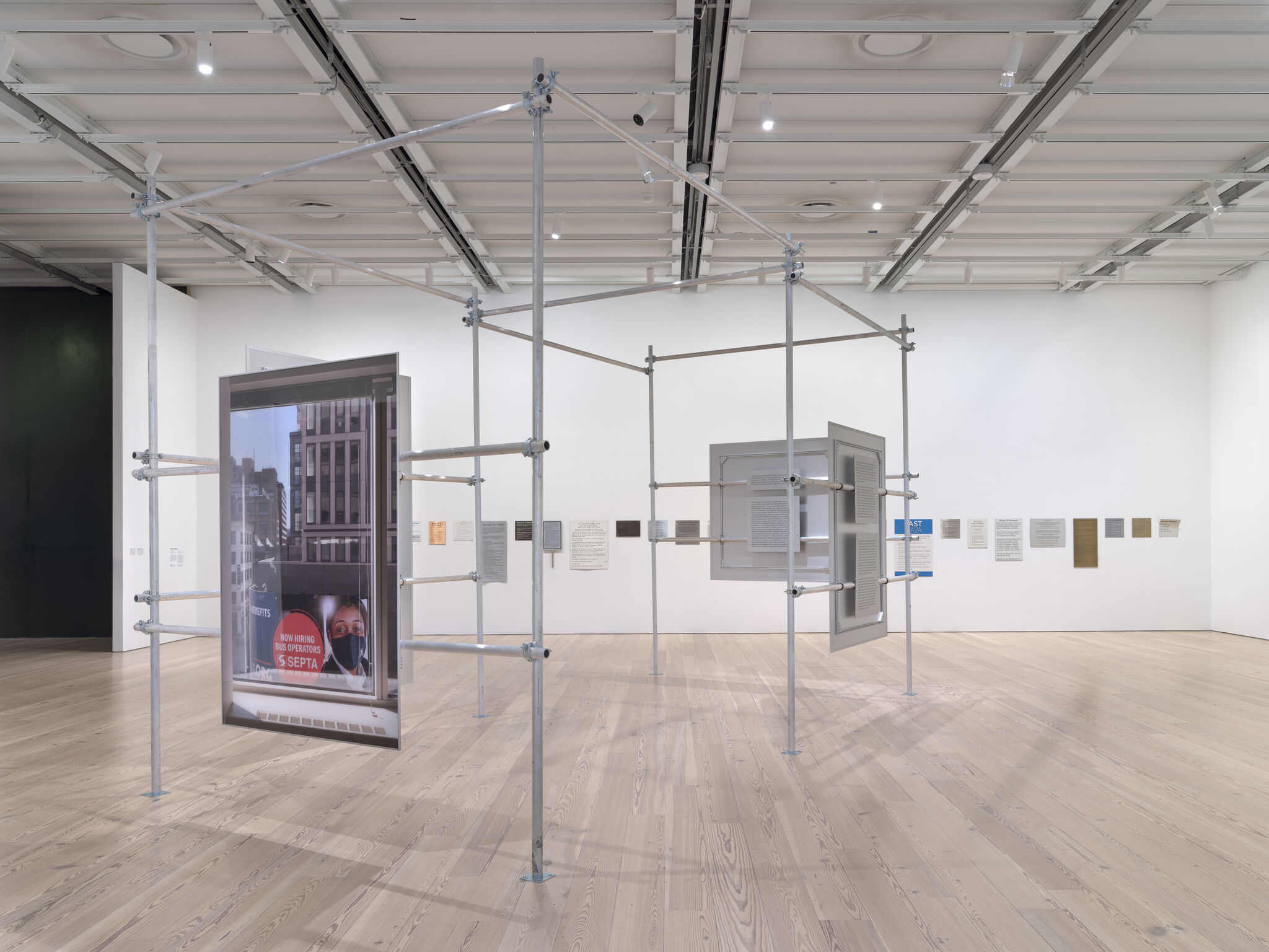 A metal scaffolding structure in a gallery displays suspended framed photographs and panels.
