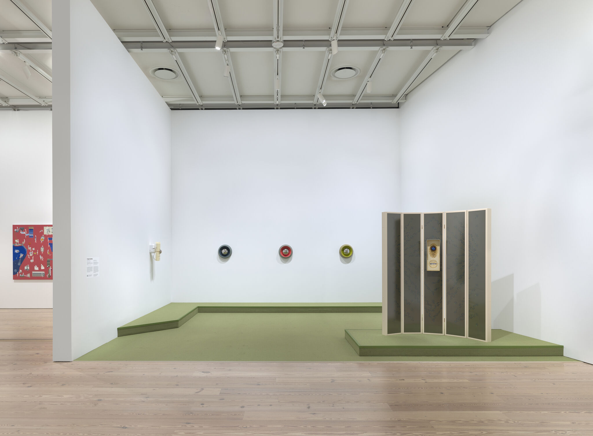 A minimalist gallery room with three colored wall sculptures and a folding screen on green platforms.