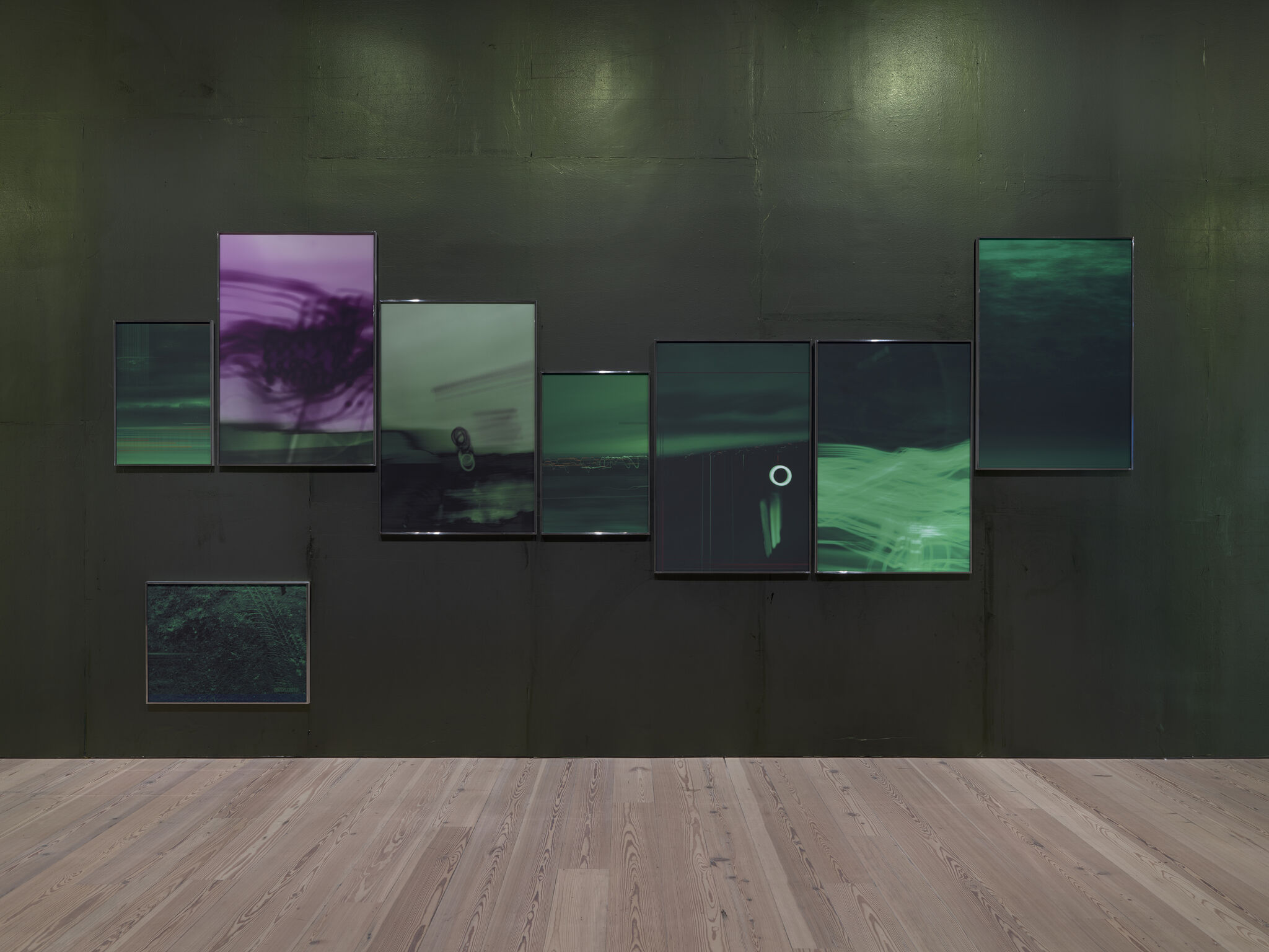 A dark gallery wall with nine framed abstract green and purple digital images arranged horizontally.
