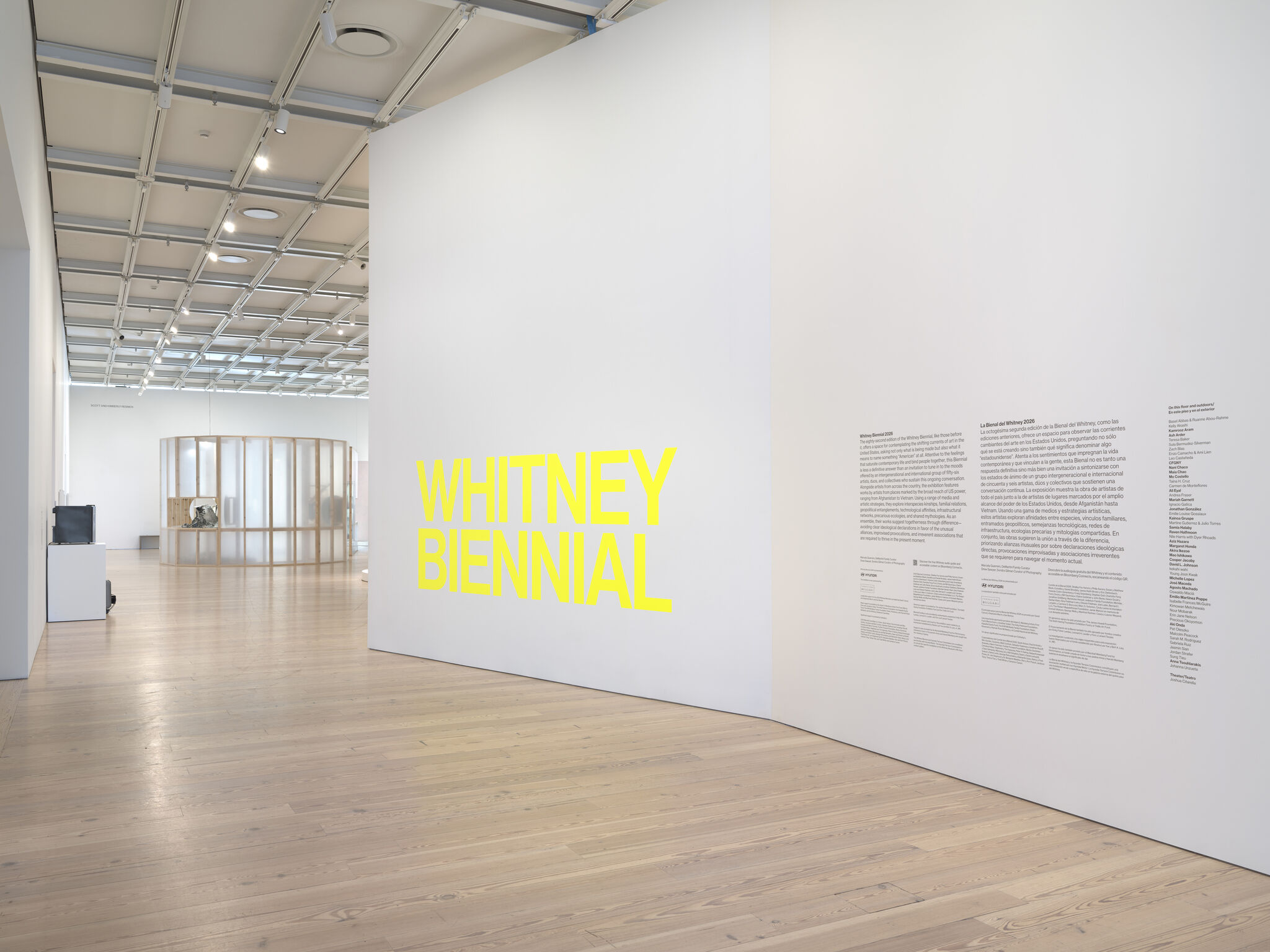 Empty gallery with a large white wall featuring bold yellow 'Whitney Biennial' text.