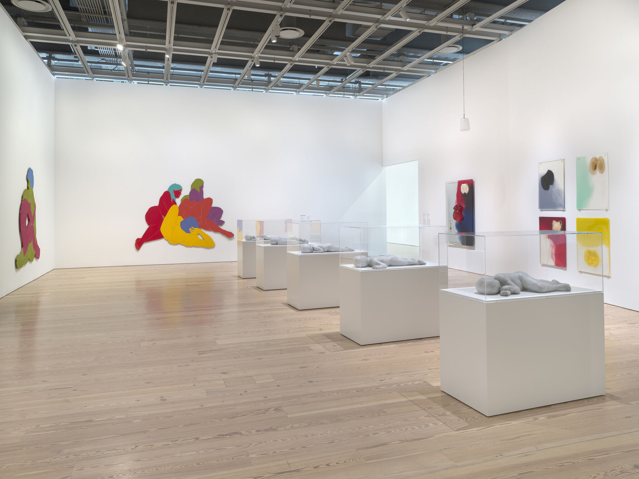 Several small reclining stone sculptures sit under glass cases amid colorful wall cutout paintings in a gallery.
