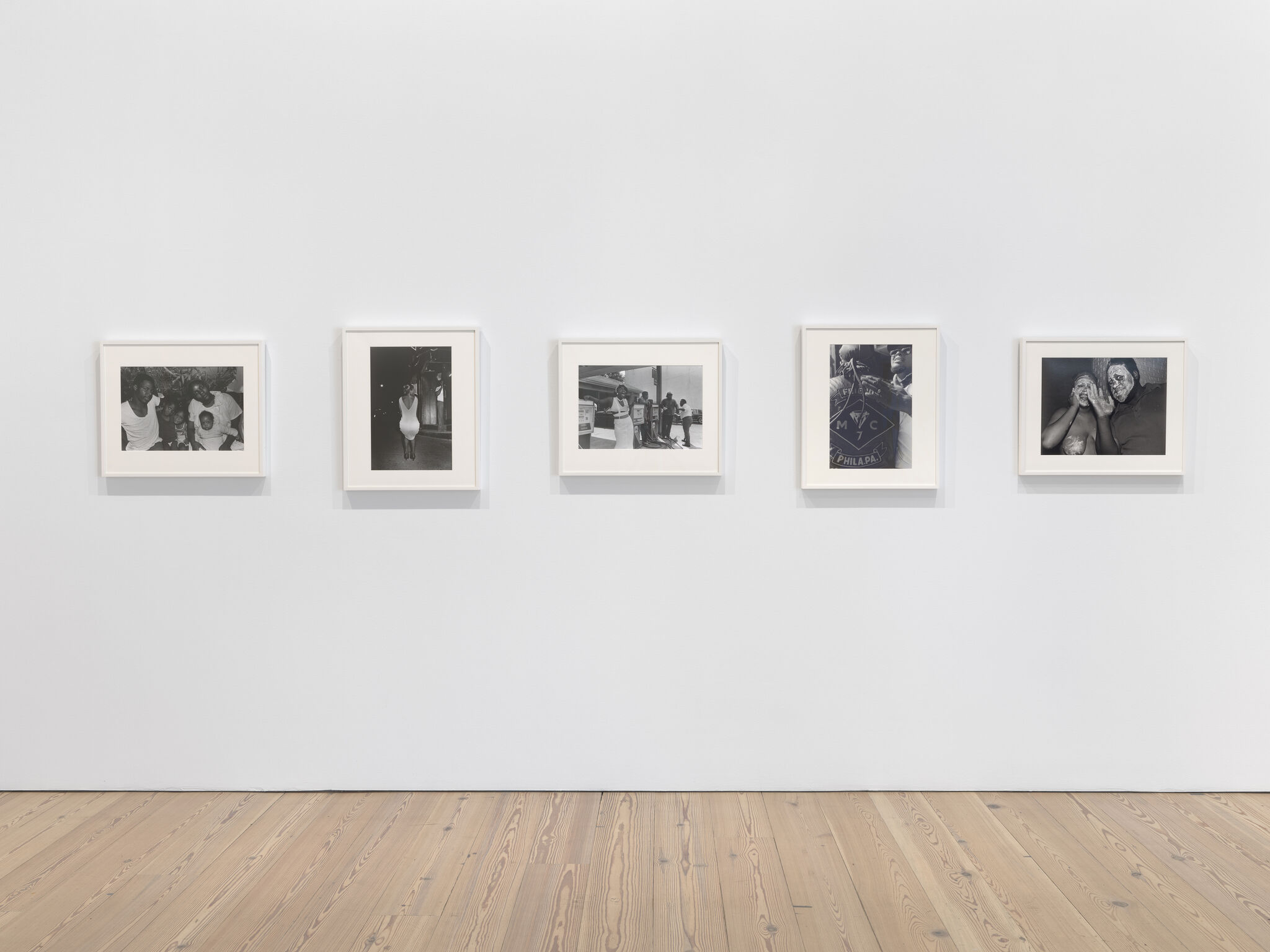 Five framed black-and-white photographs hang in a row on a white gallery wall above wooden floor.