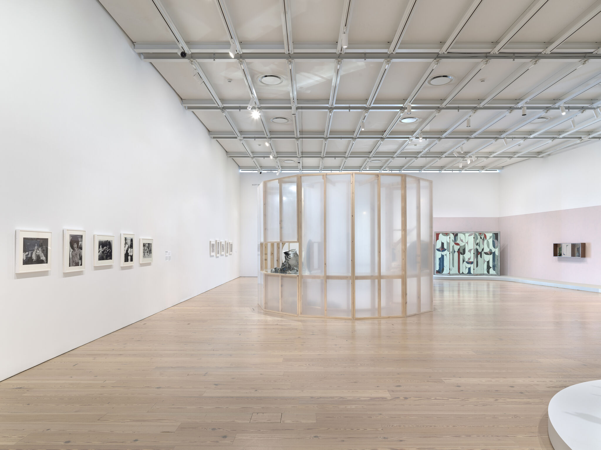 A translucent wooden curved partition encloses sculptures in a bright, minimal contemporary art gallery room.