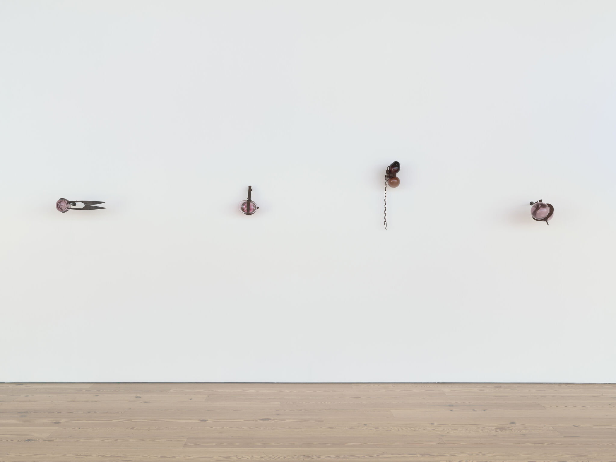 Four small pink glass and metal sculptures mounted in a row on a white gallery wall.