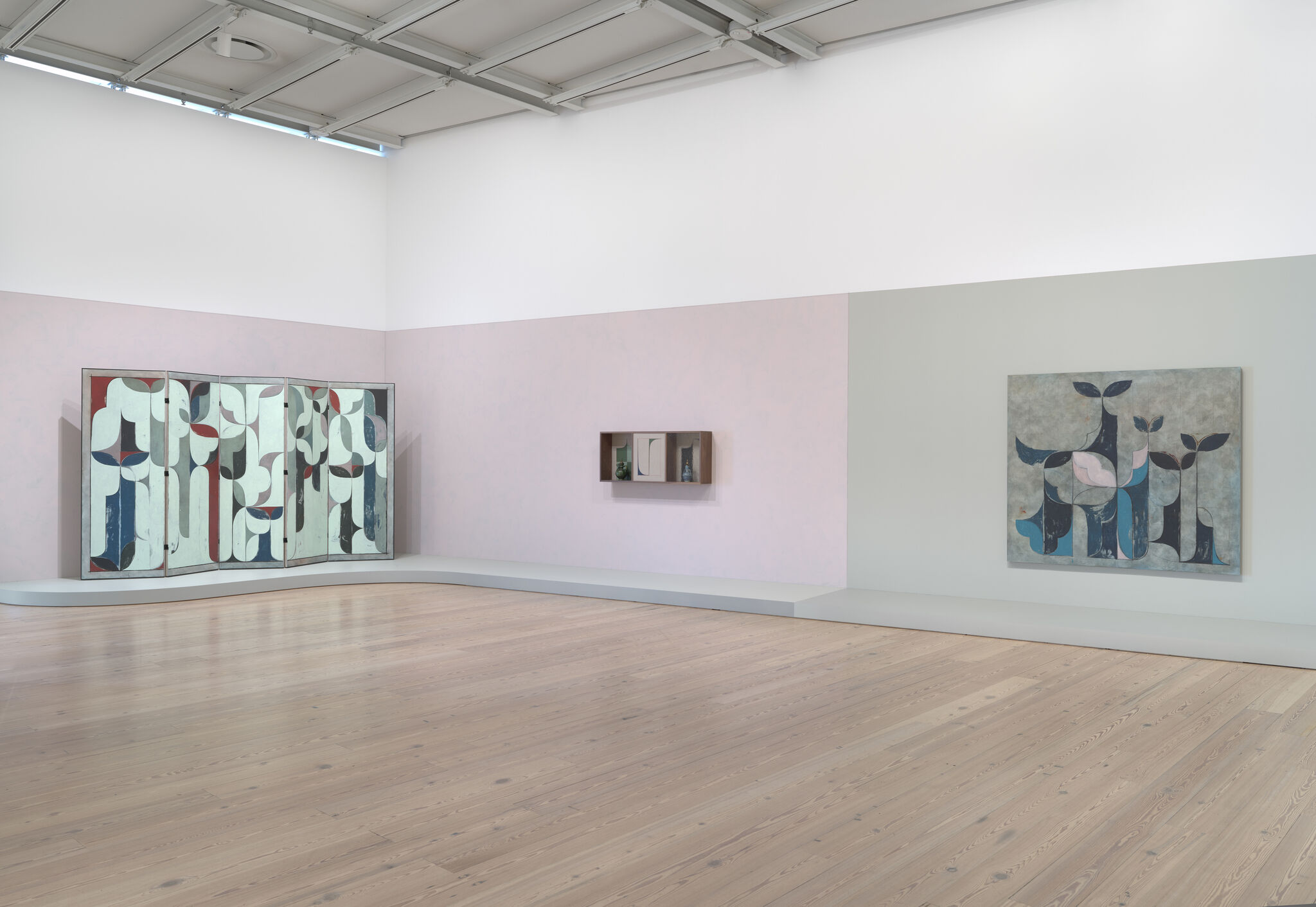 Gallery room with abstract blue and pink paintings displayed on pale walls above a wooden floor.