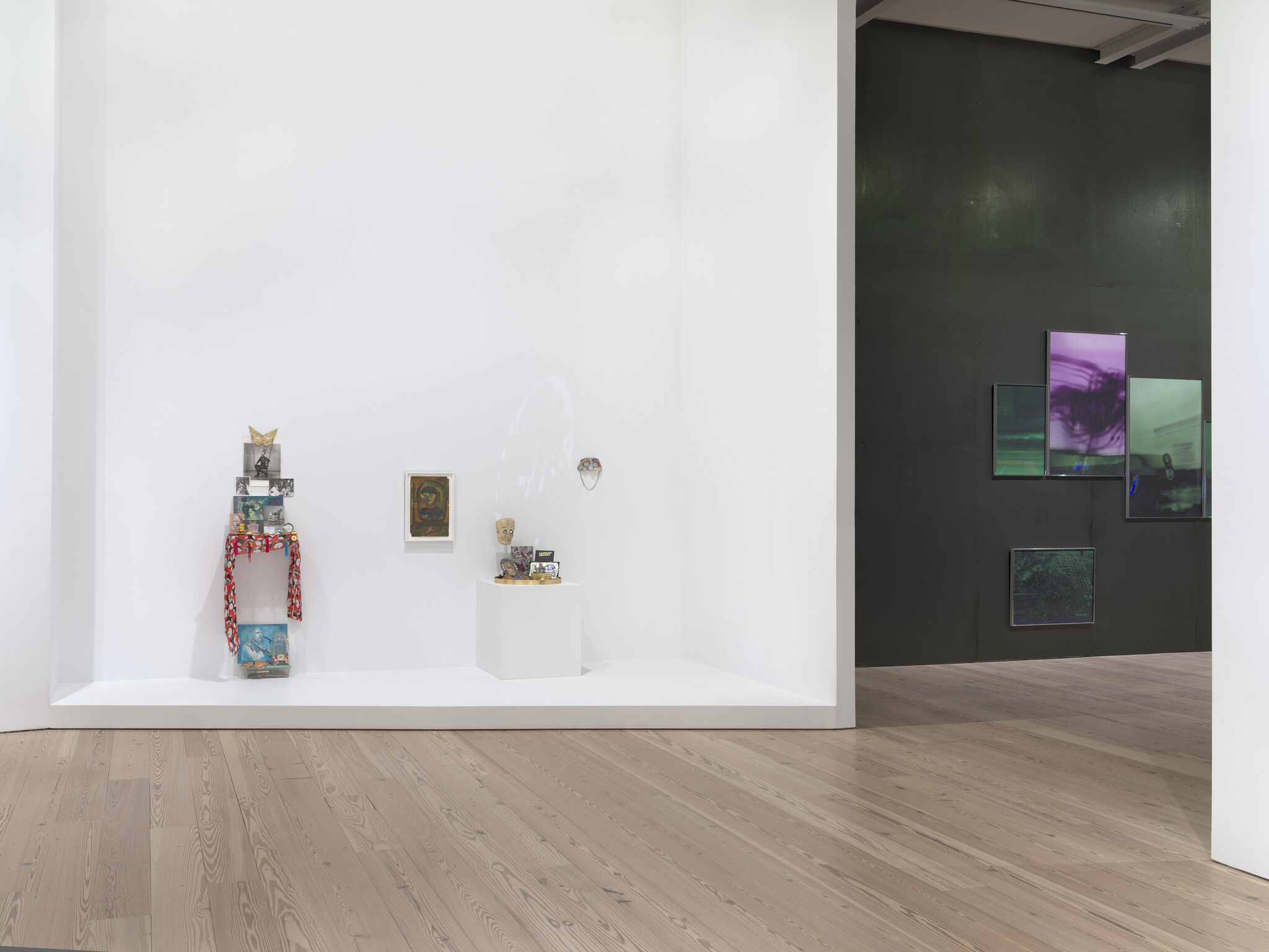 Small mixed-media sculptures and framed pictures displayed on a white gallery wall beside colorful digital screens.