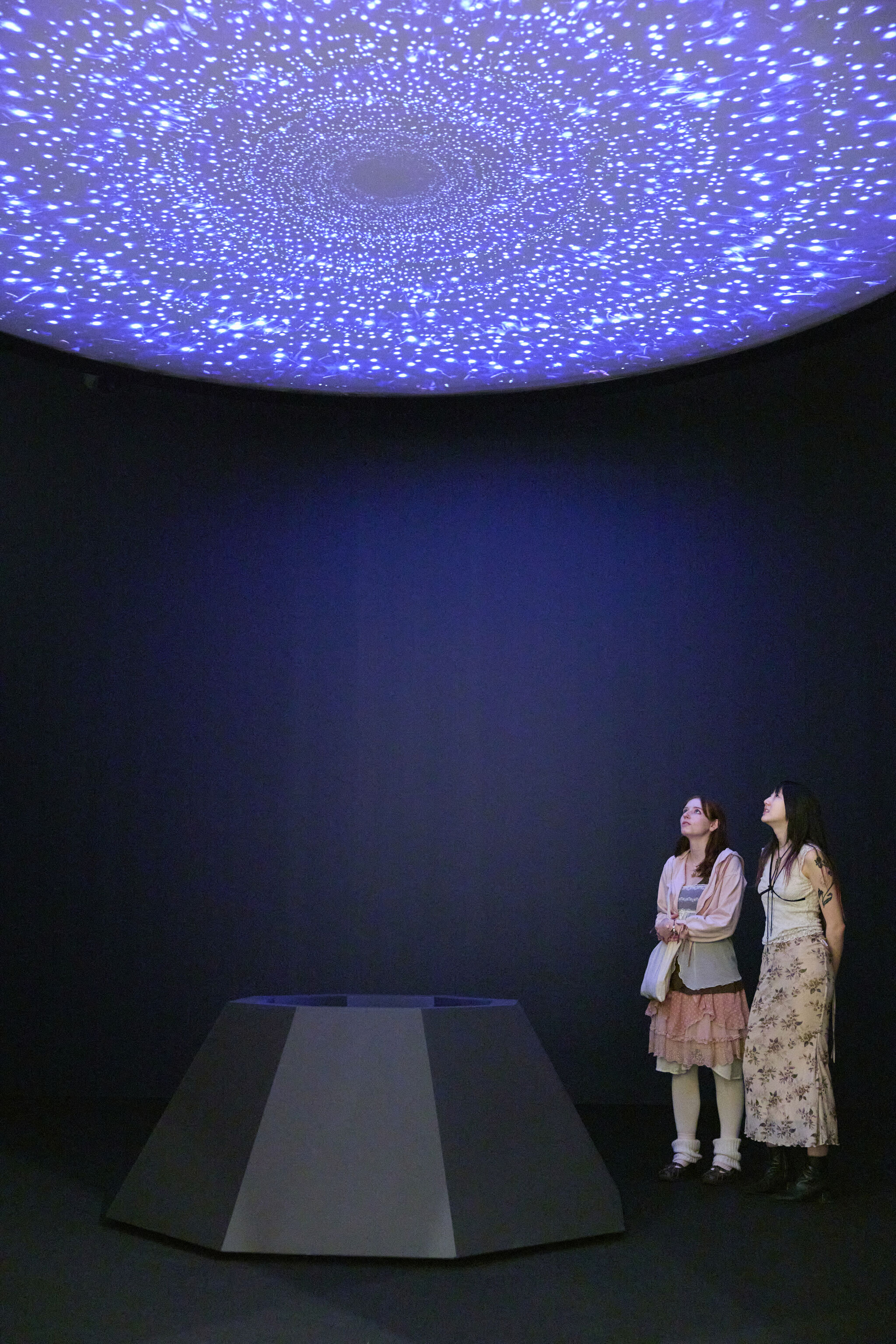 Two women stand and gaze upward at a blue, star-like ceiling projection in a dark room.
