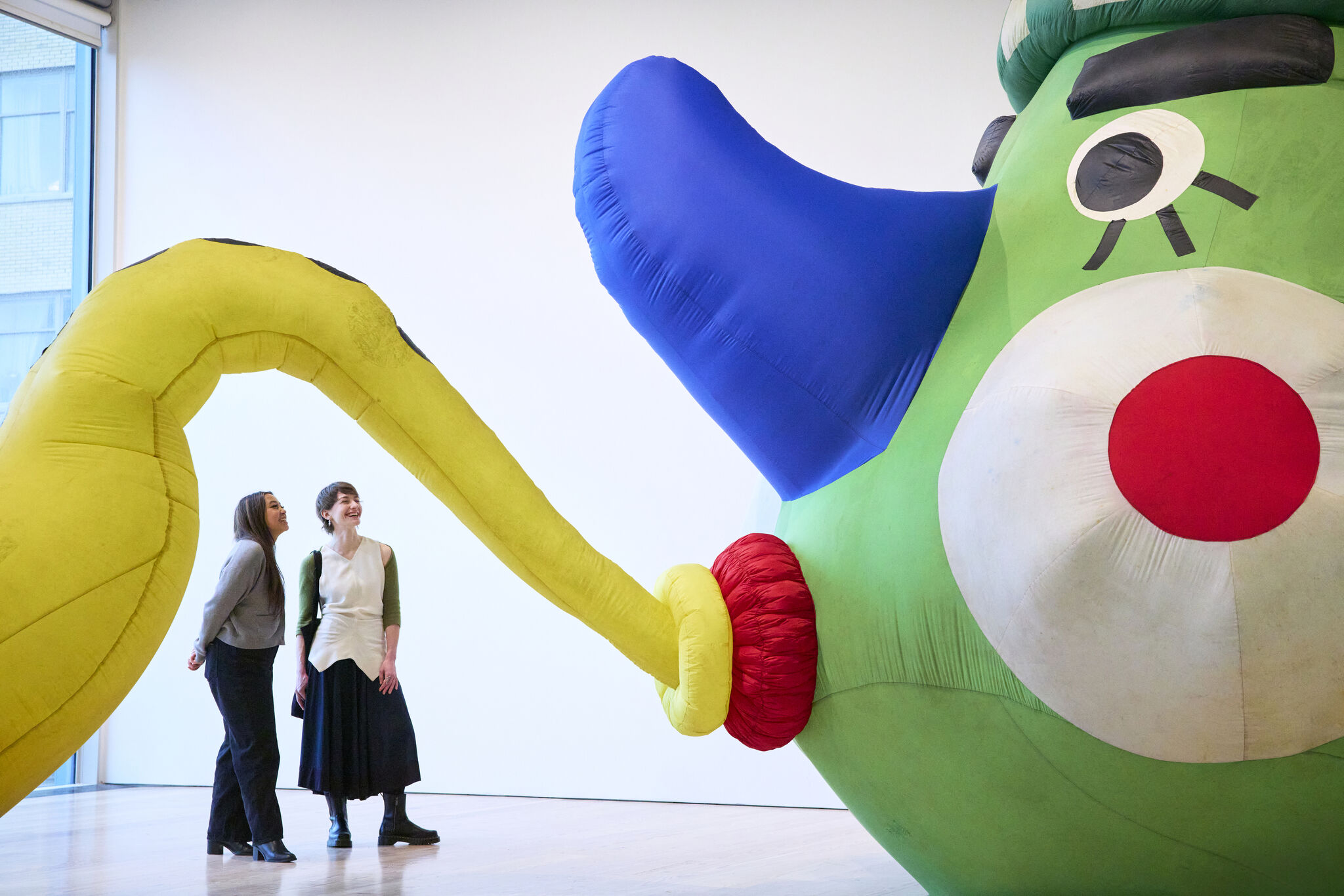 Two women stand smiling while looking at a giant colorful inflatable sculpture with a long yellow tube.