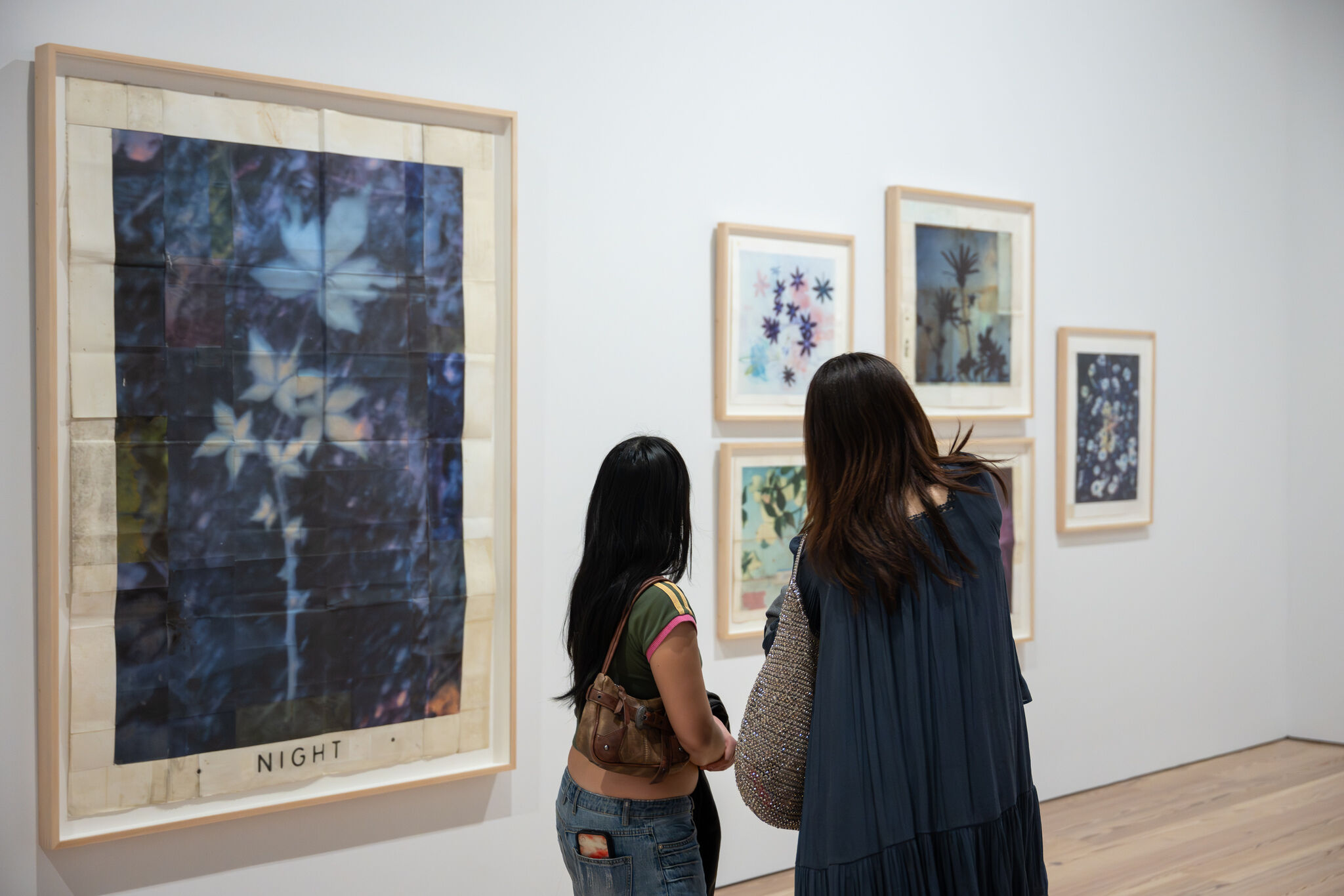 Two people closely examine framed botanical artworks on a gallery wall, including a large piece labeled Night.