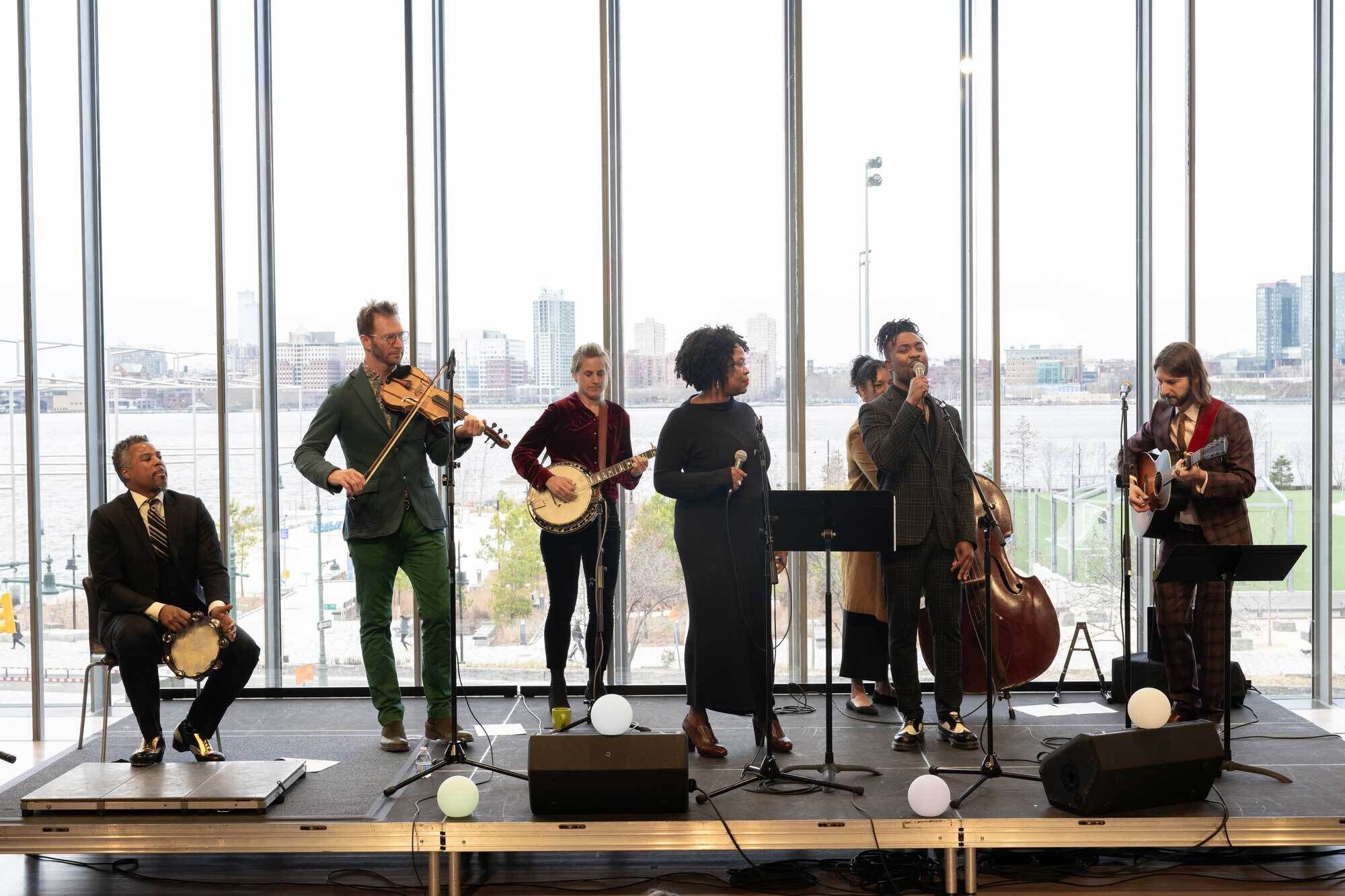 A group of Jazz at Lincoln Center musicians perform on a small stage inside the Whitney Museum, framed by tall floor-to-ceiling windows overlooking the city and waterfront. The ensemble includes singers and instrumentalists playing violin, banjo, guitar, upright bass, and percussion, creating a lively, collaborative performance. 