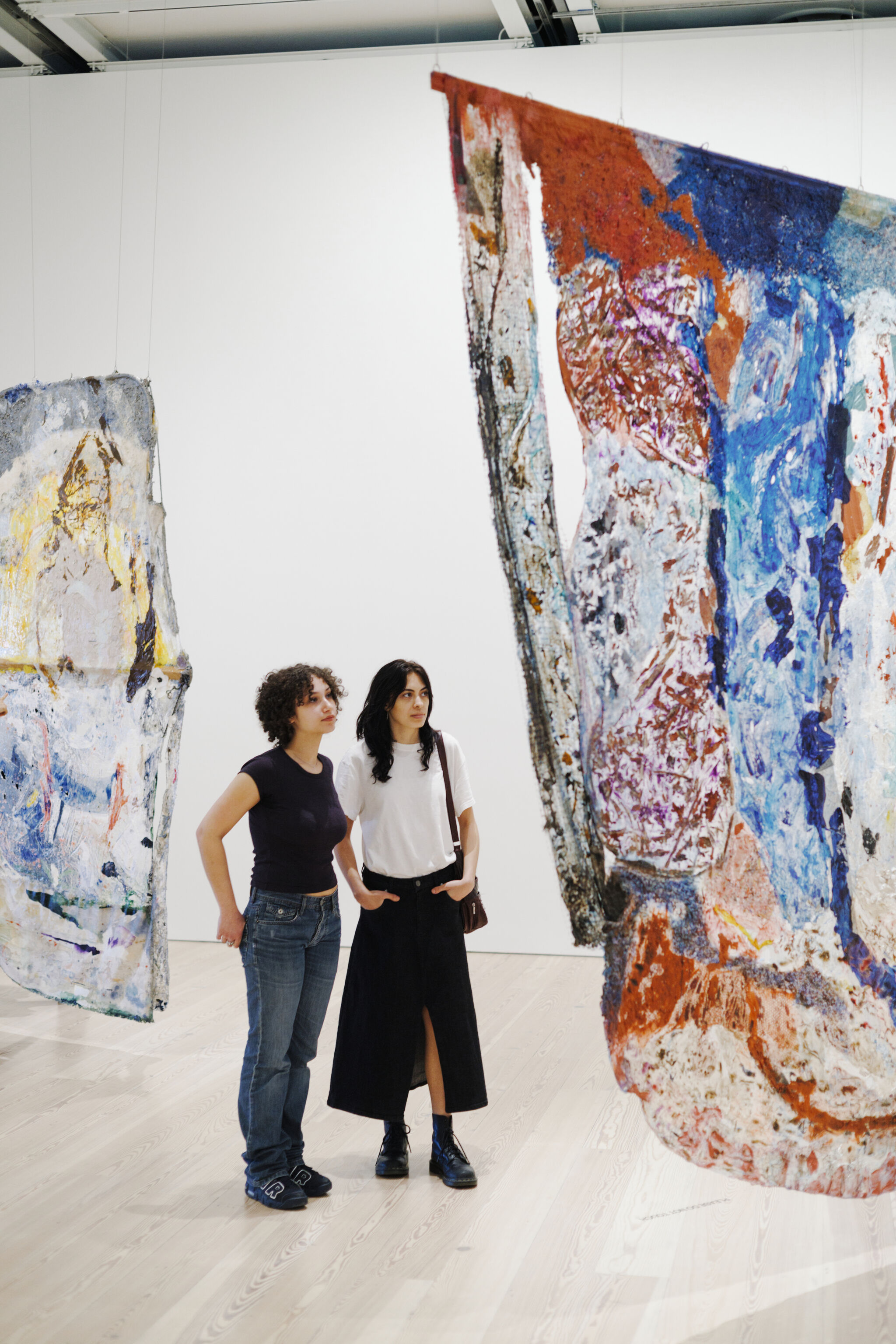 Two women stand in a gallery looking attentively at large colorful hanging abstract paintings.