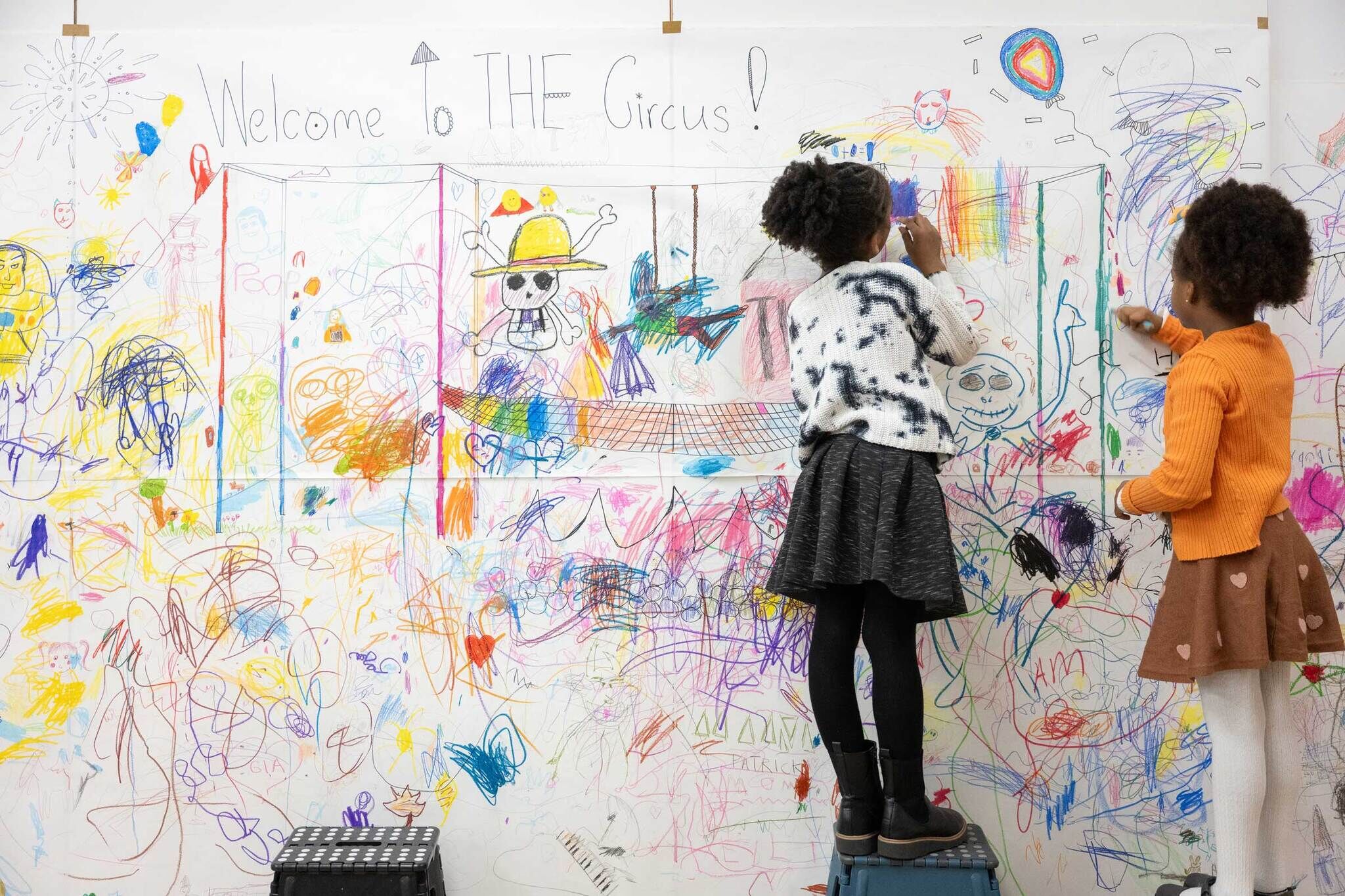 Two young children stand on small stools, drawing with colorful markers on a large wall-sized paper mural filled with playful scribbles, shapes, and figures. The mural includes the handwritten phrase “Welcome to THE Circus!” and is covered with layered, vibrant marks and imaginative drawings. 
