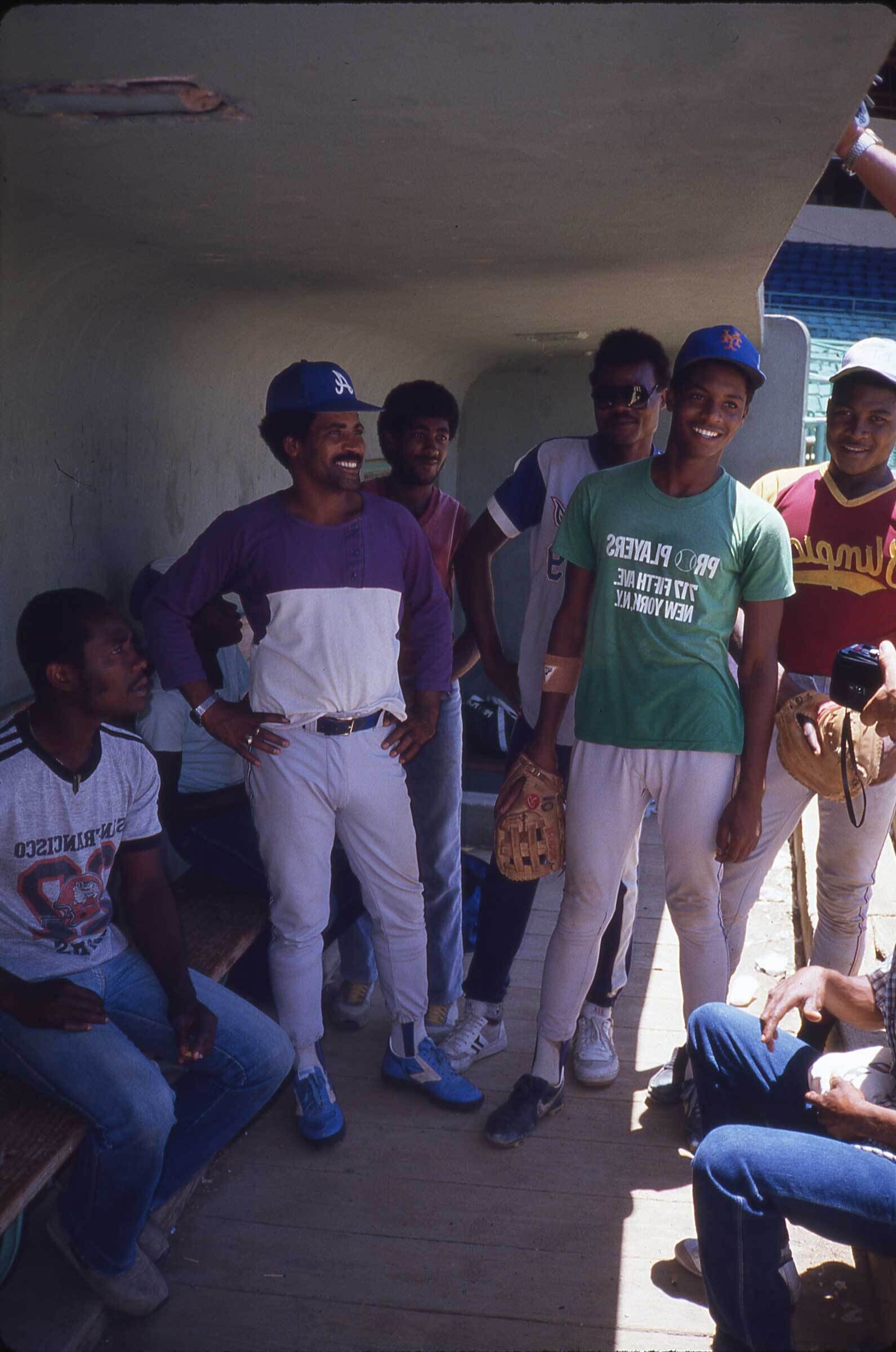 Young baseball players stand and sit in a dugout, smiling and holding baseball gloves.