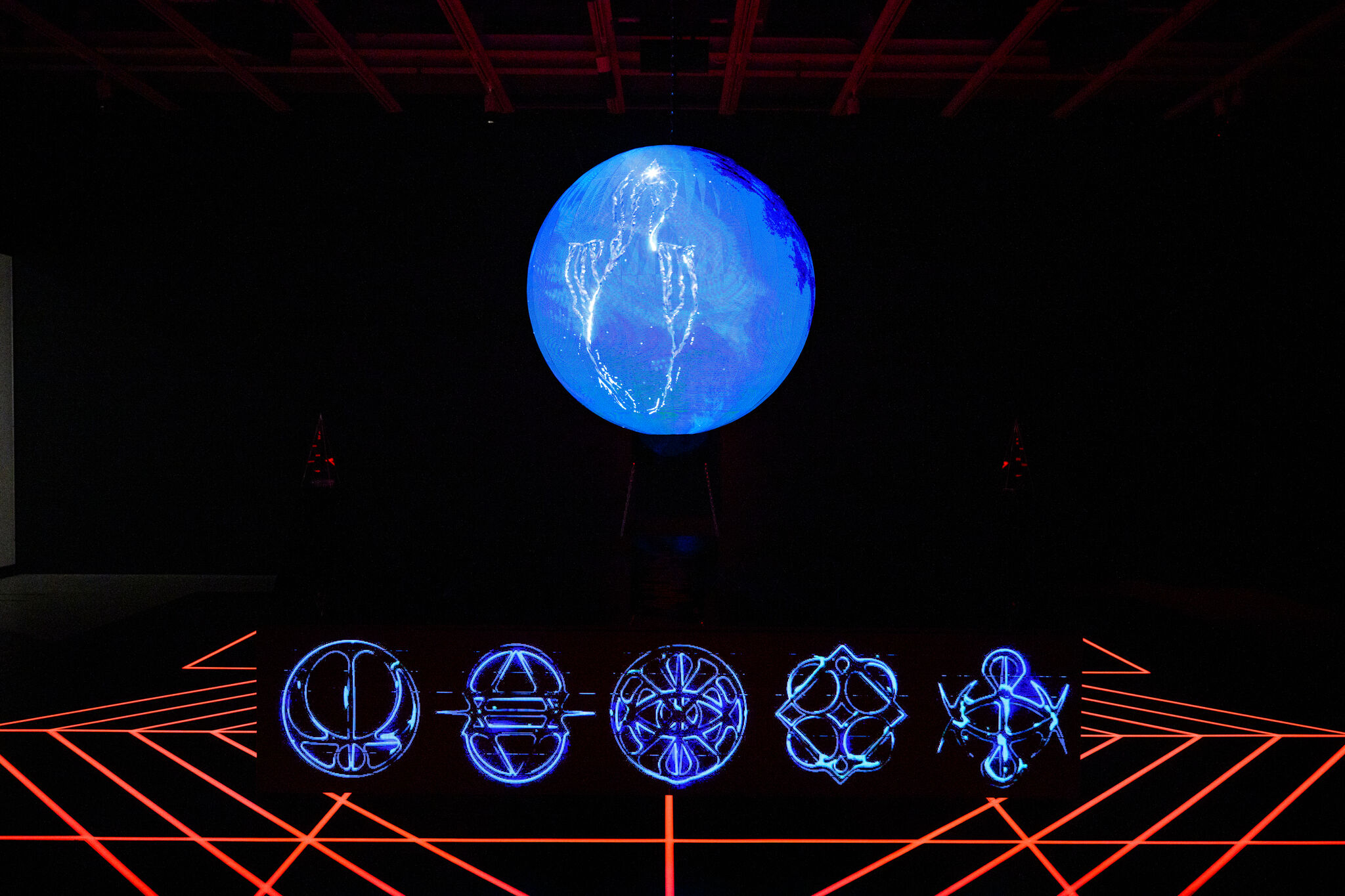 View of Zac Blas' installation, Cultus, featuring a dark gallery space with a large glowing blue sphere suspended above five neon-blue circular symbols arranged in a row on top of a black floor crossed by red laser grid lines.