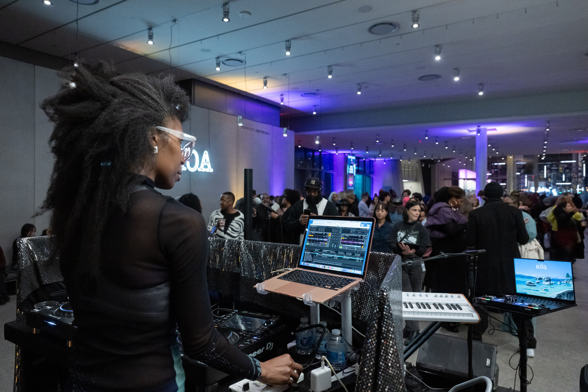 A DJ adjusts controls at a laptop and turntables while a crowd watches in purple-lit venue.