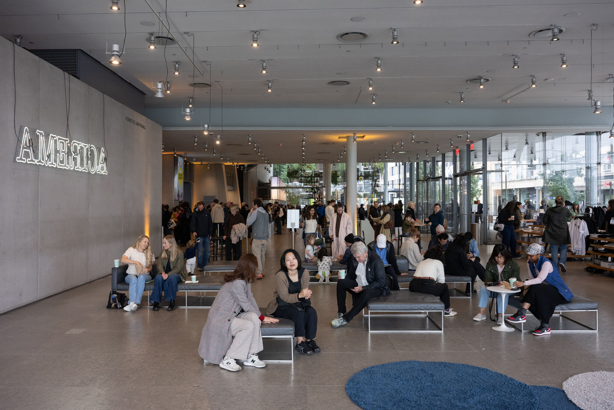 People sit, talk, and use devices in a busy modern museum lobby with large windows.