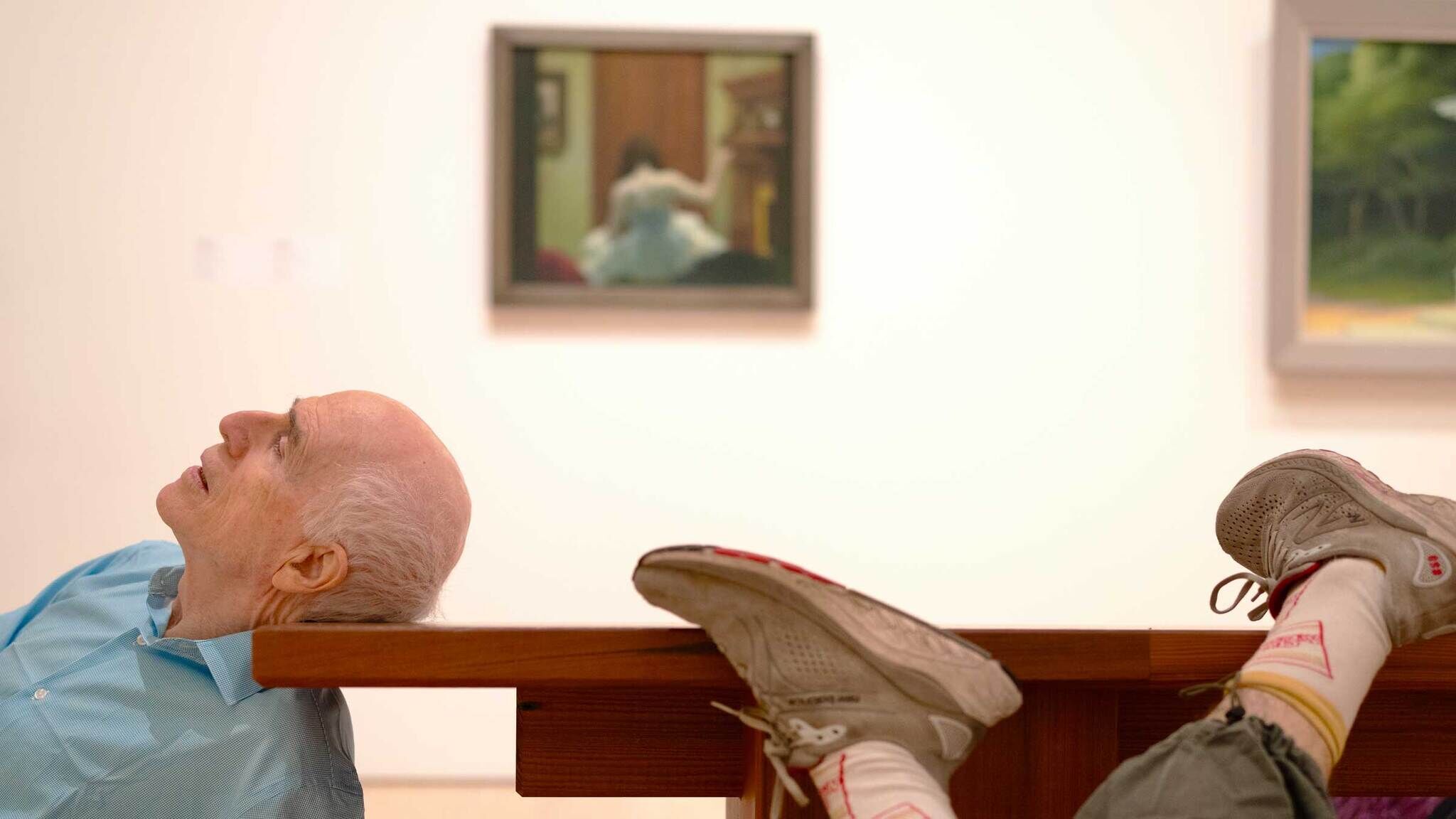 An elderly man leans back on a gallery bench looking up while someone rests their feet.
