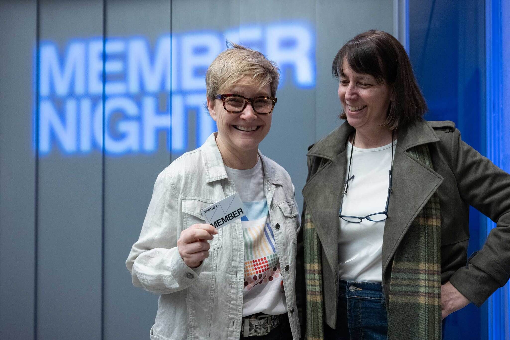 Two Whitney Members stand in front of the Museum's elevator with the words Member Night projected onto it. One member holds up a Membership card.