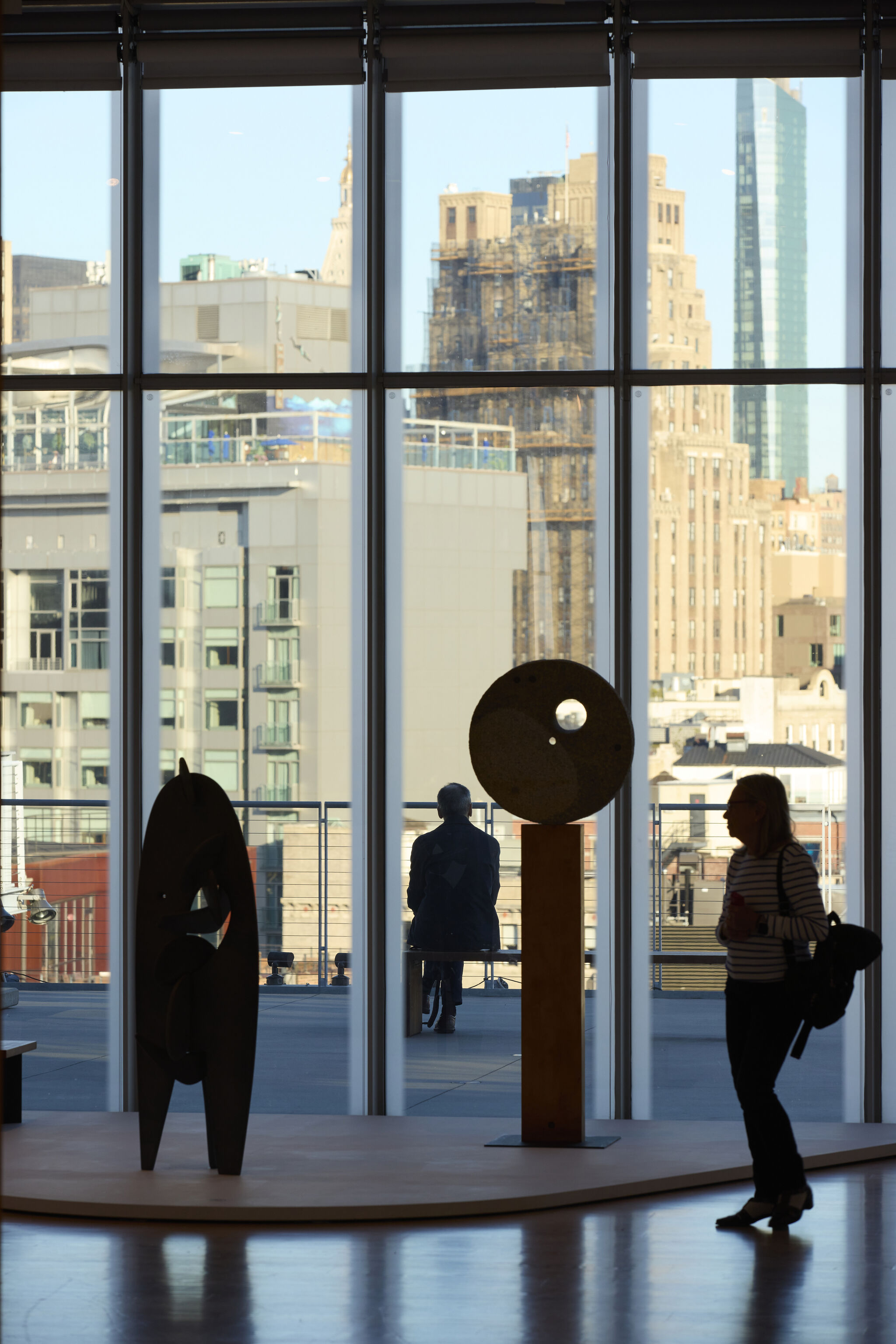In front of floor to ceiling windows, two sculptures on a raised platform are encased by a large dark shadow. In the foreground, a woman is walking by and in the background on the terrace outside the window, a man is sitting down on a bench overlooking the city.