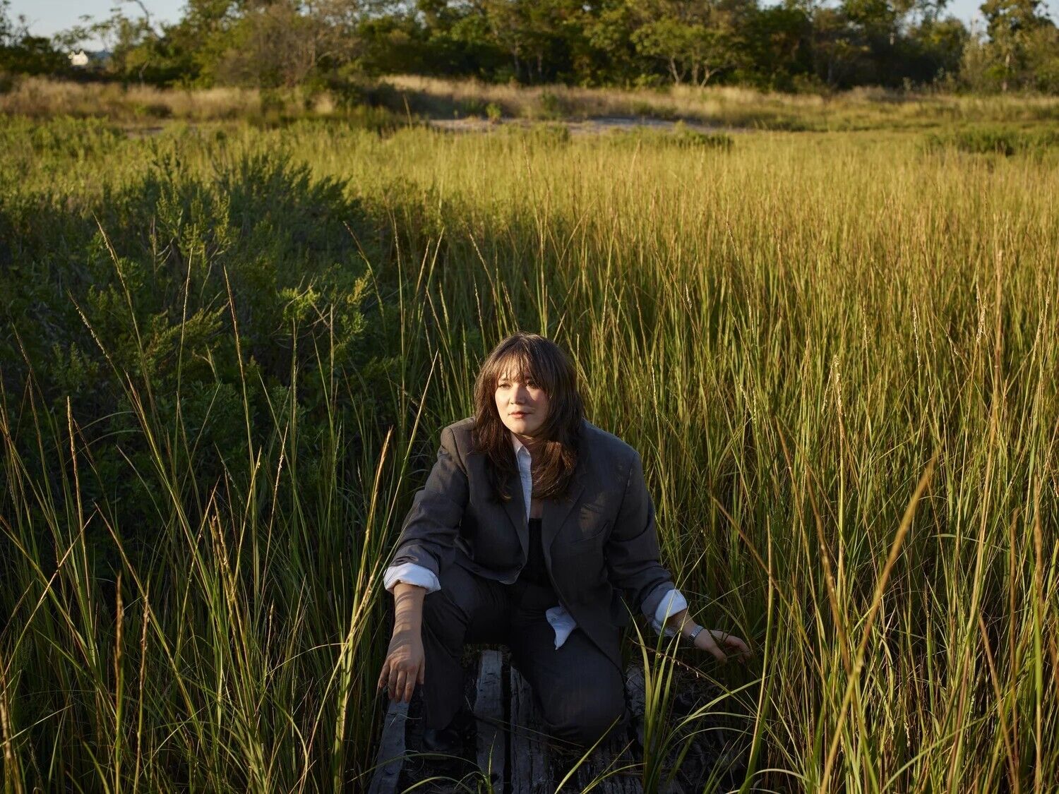 Wendy Eisenberg, a woman with long brown hair in a grey suit is posing in a grassy field.