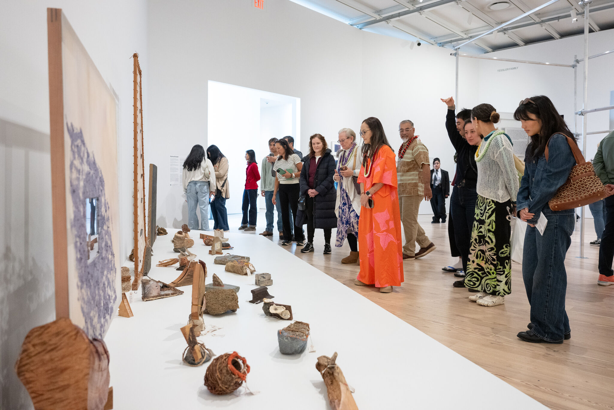A diverse group of visitors attentively examines small artifacts displayed on a white gallery table.