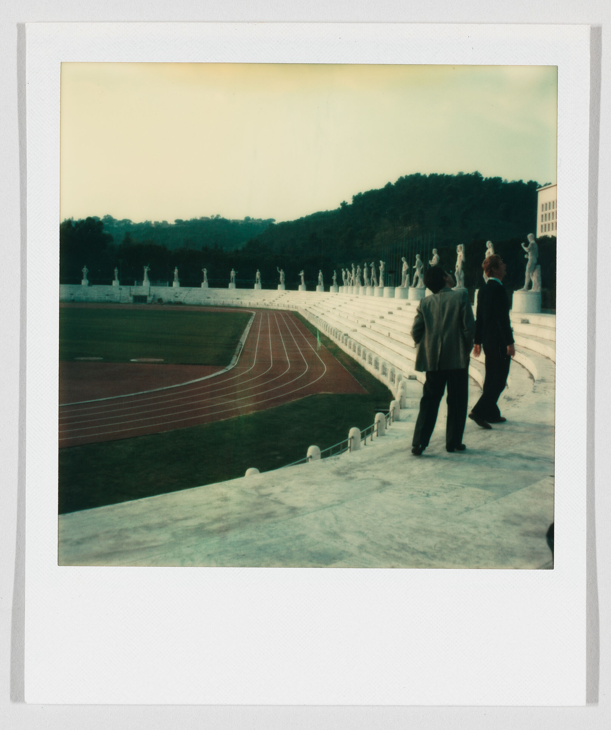 Two men in suits walk along marble stadium steps past a row of classical statues.