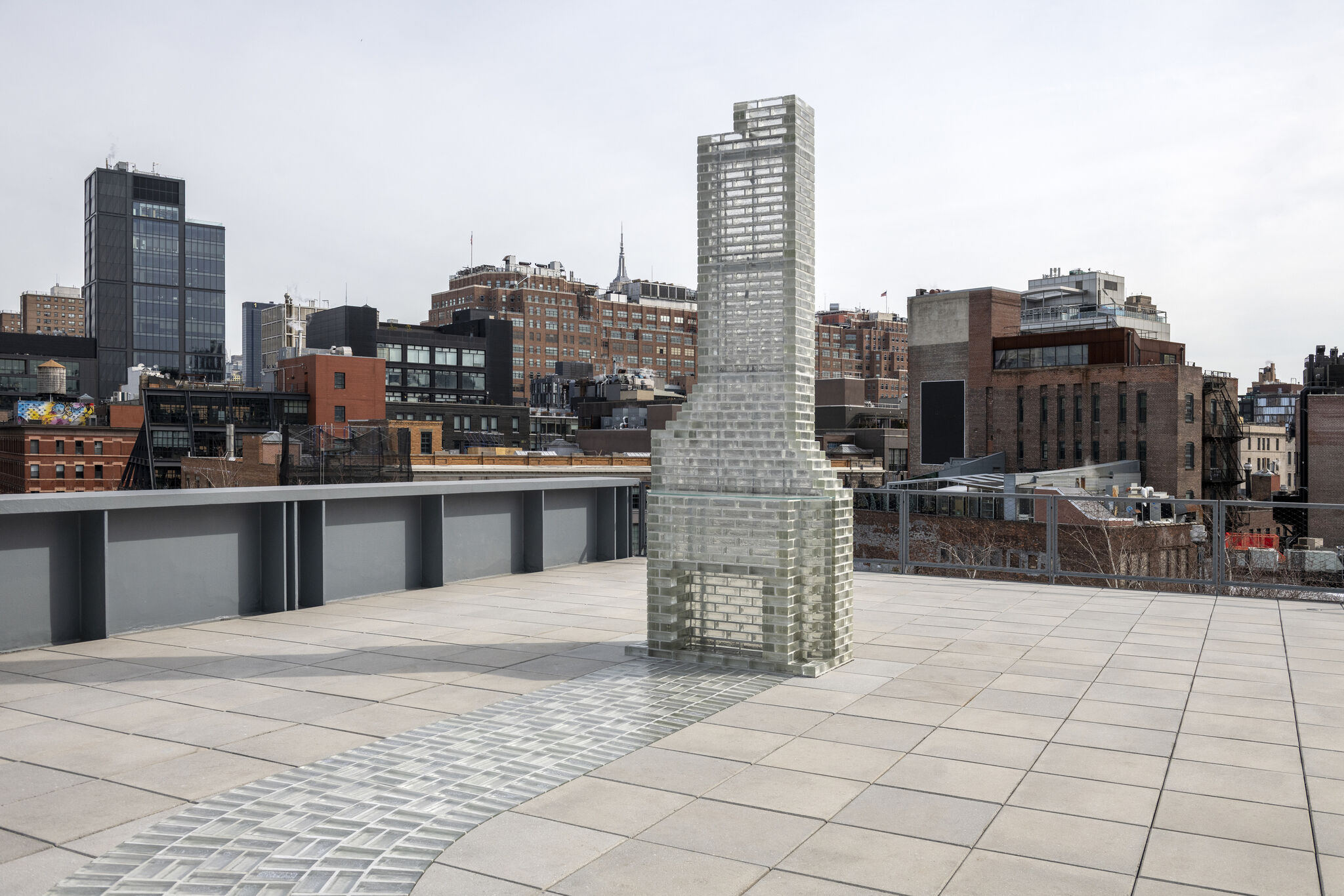Glass brick tower sculpture stands on a rooftop terrace with city buildings in the background.