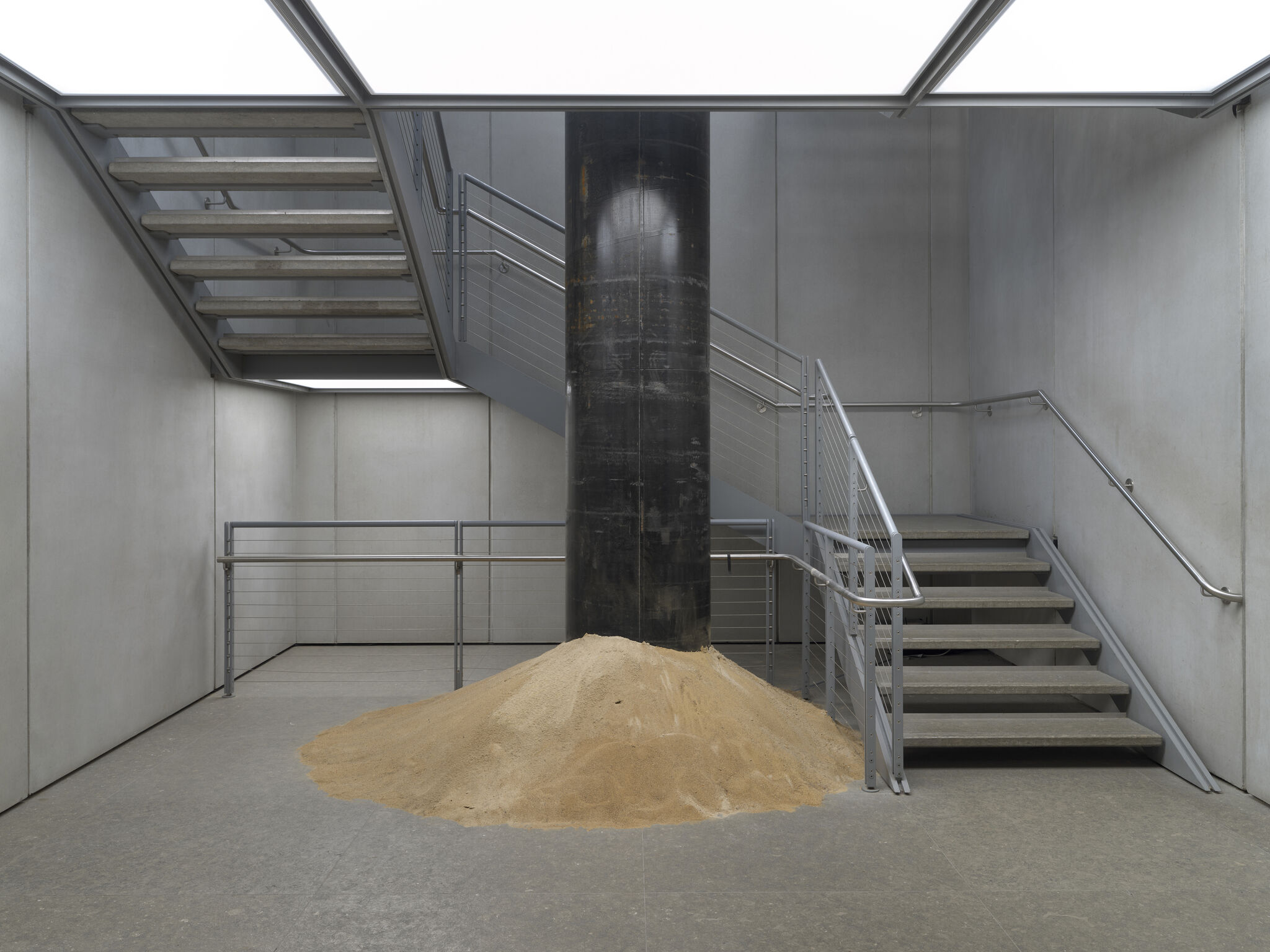 A pile of sand sits beneath a large black pipe in a gray concrete stairwell.