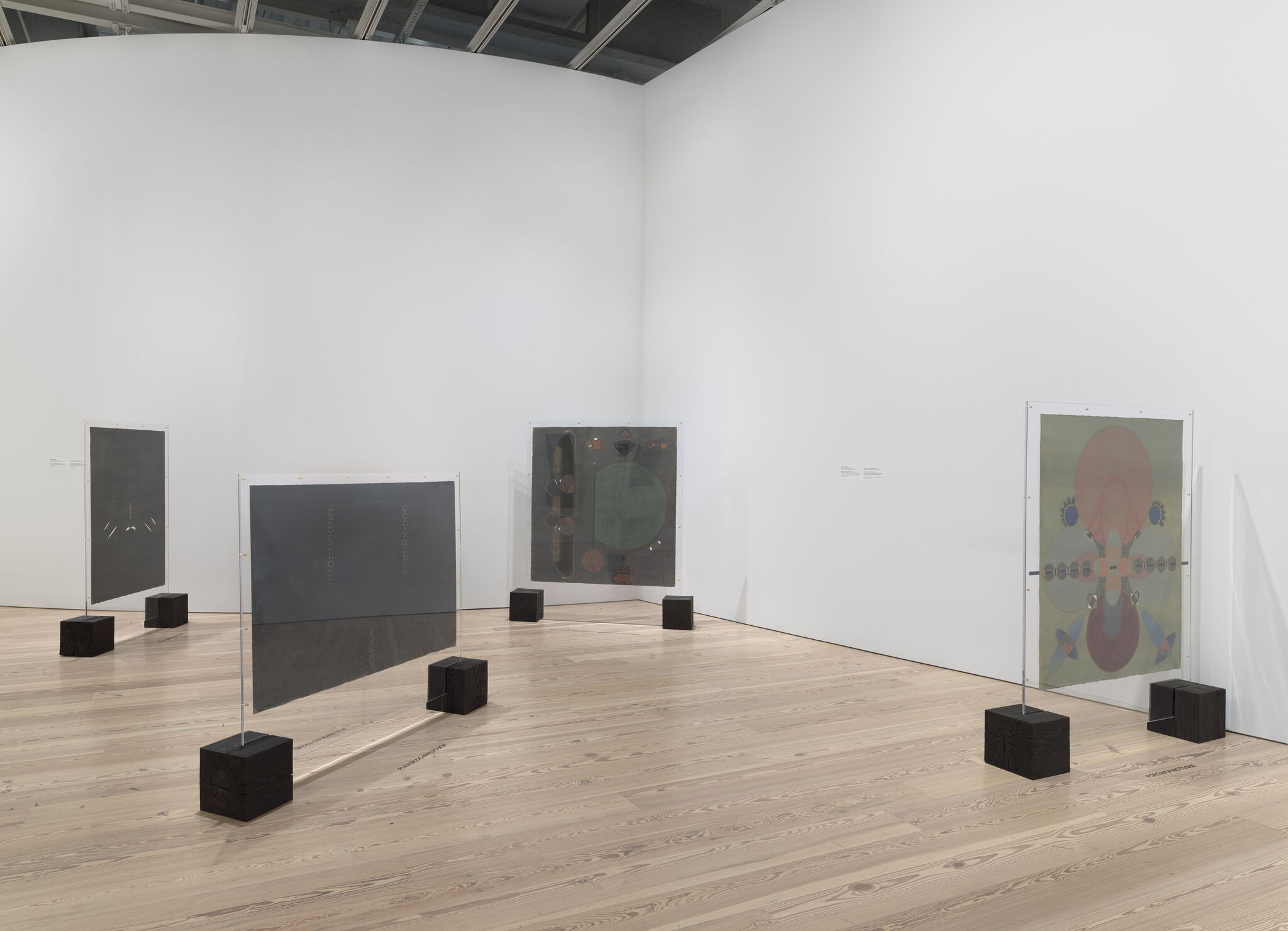 Several translucent panels and framed artworks stand on wooden blocks in a bright white gallery space.