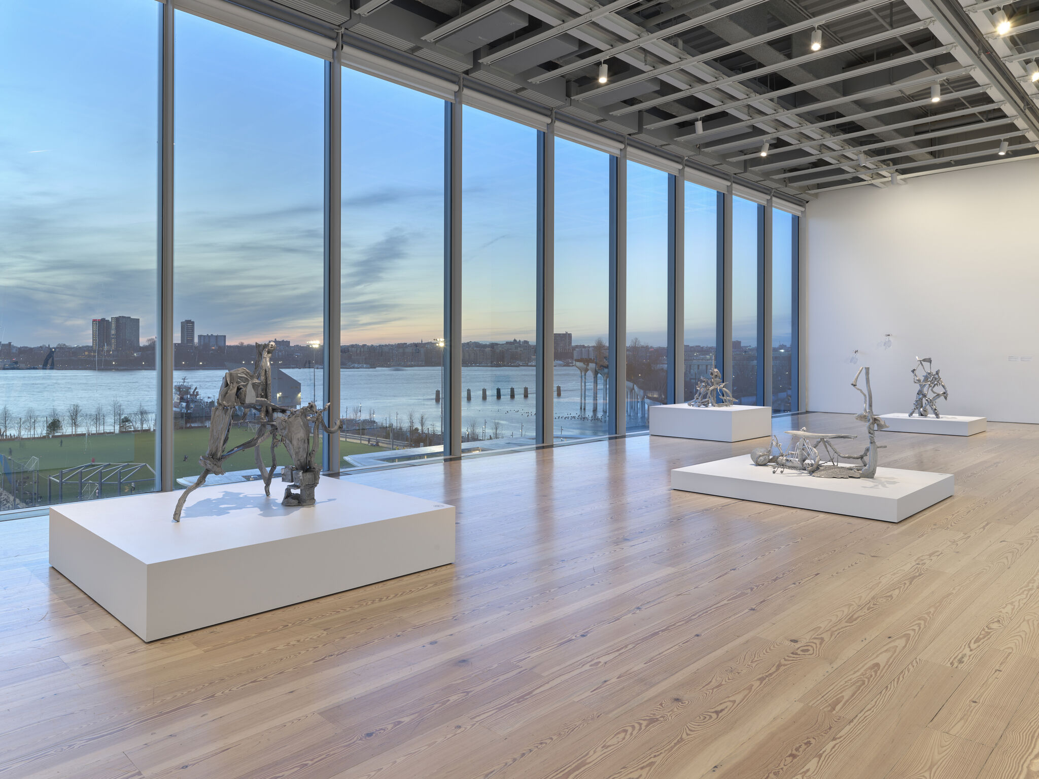 Silver abstract sculptures on white pedastals in front of big, floor-to-ceiling windows overlooking a window at sunset.