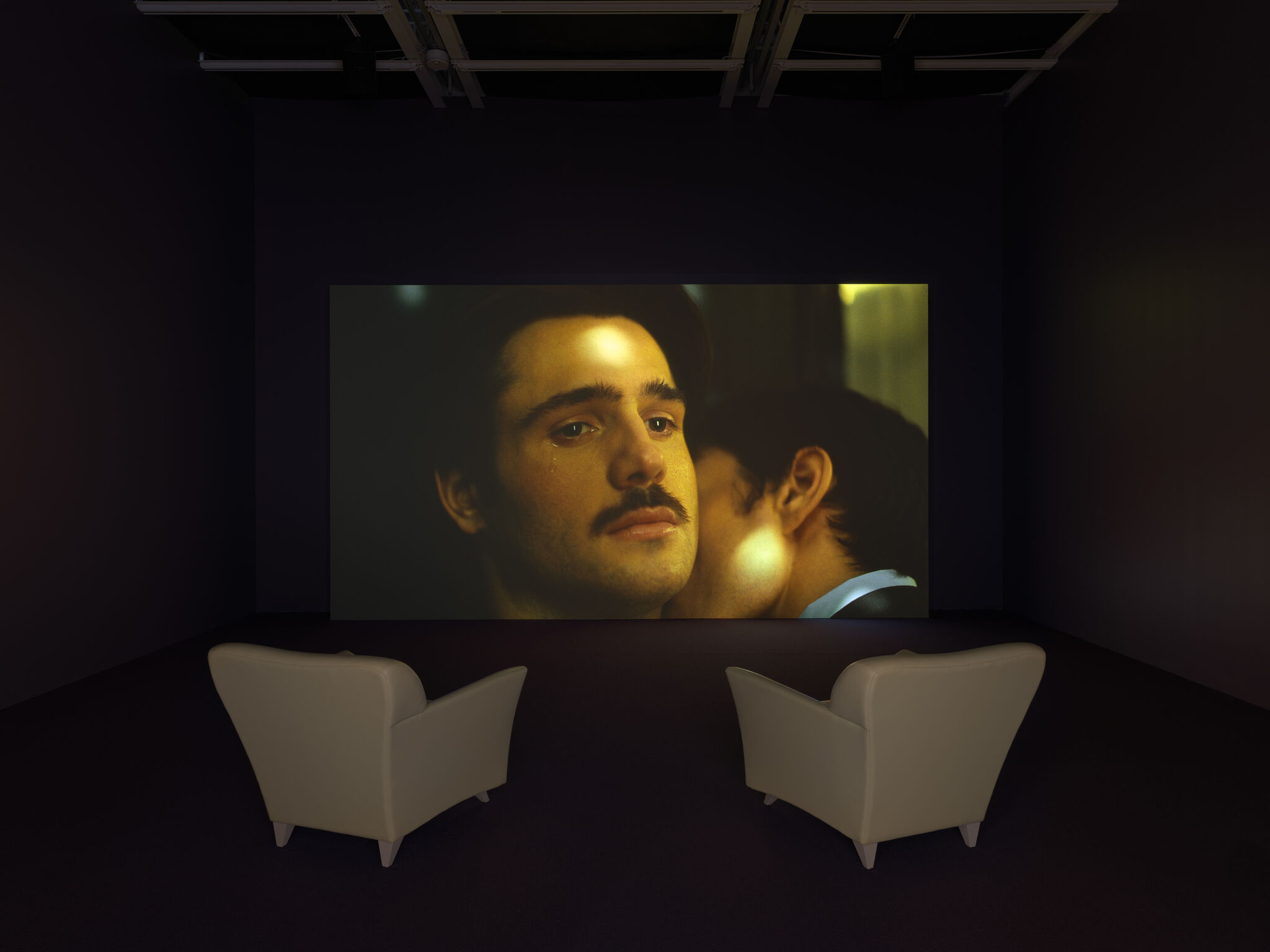 A dark room with two armchairs facing a screen. On the screen is a man looking forlornly into the distance while another man kisses his neck.