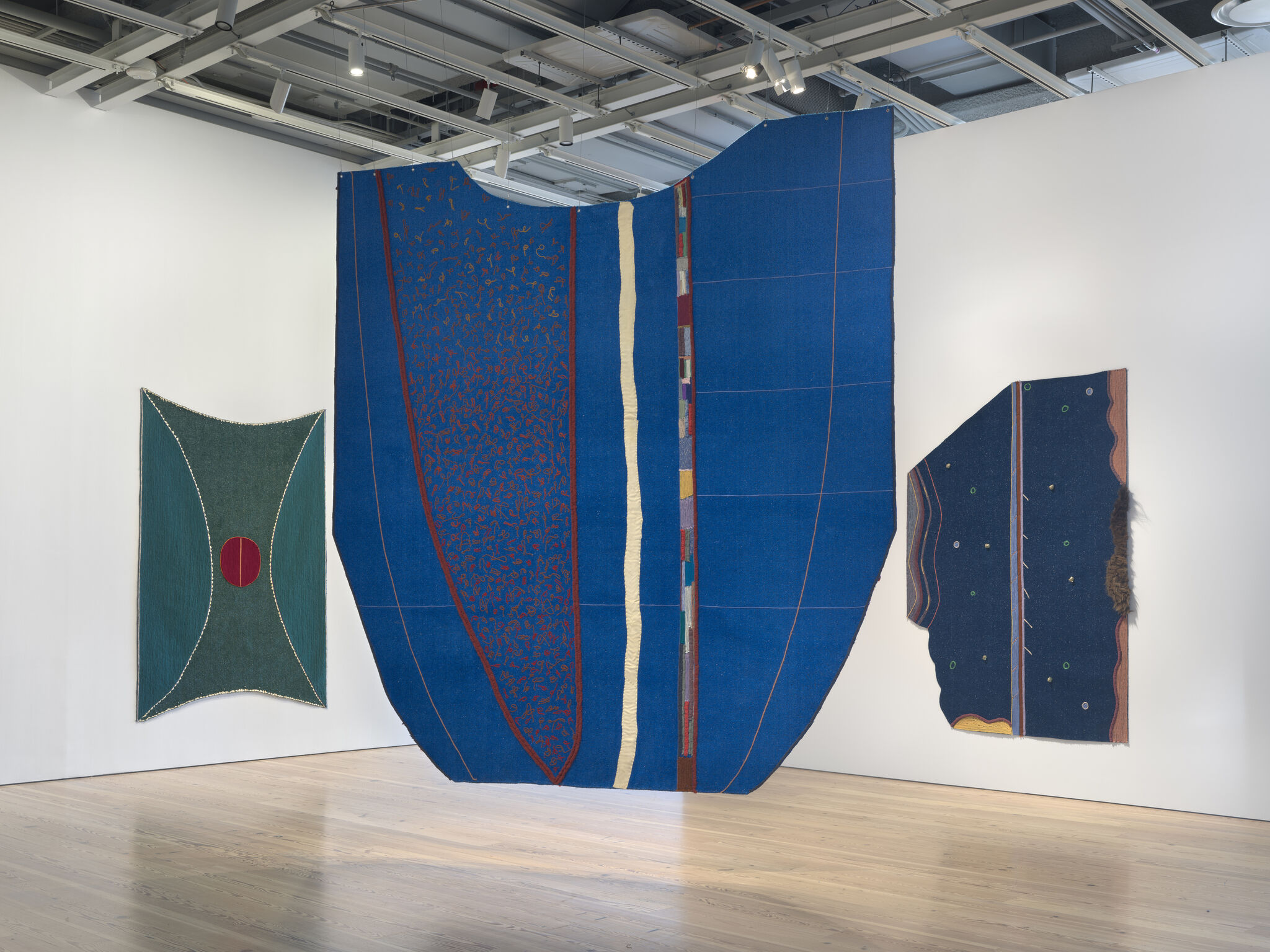 Three big fabric artwork pieces made of bright blues and greens. Two hang on walls on the left and right and in the middle one of the artworks hangs from the ceiling.