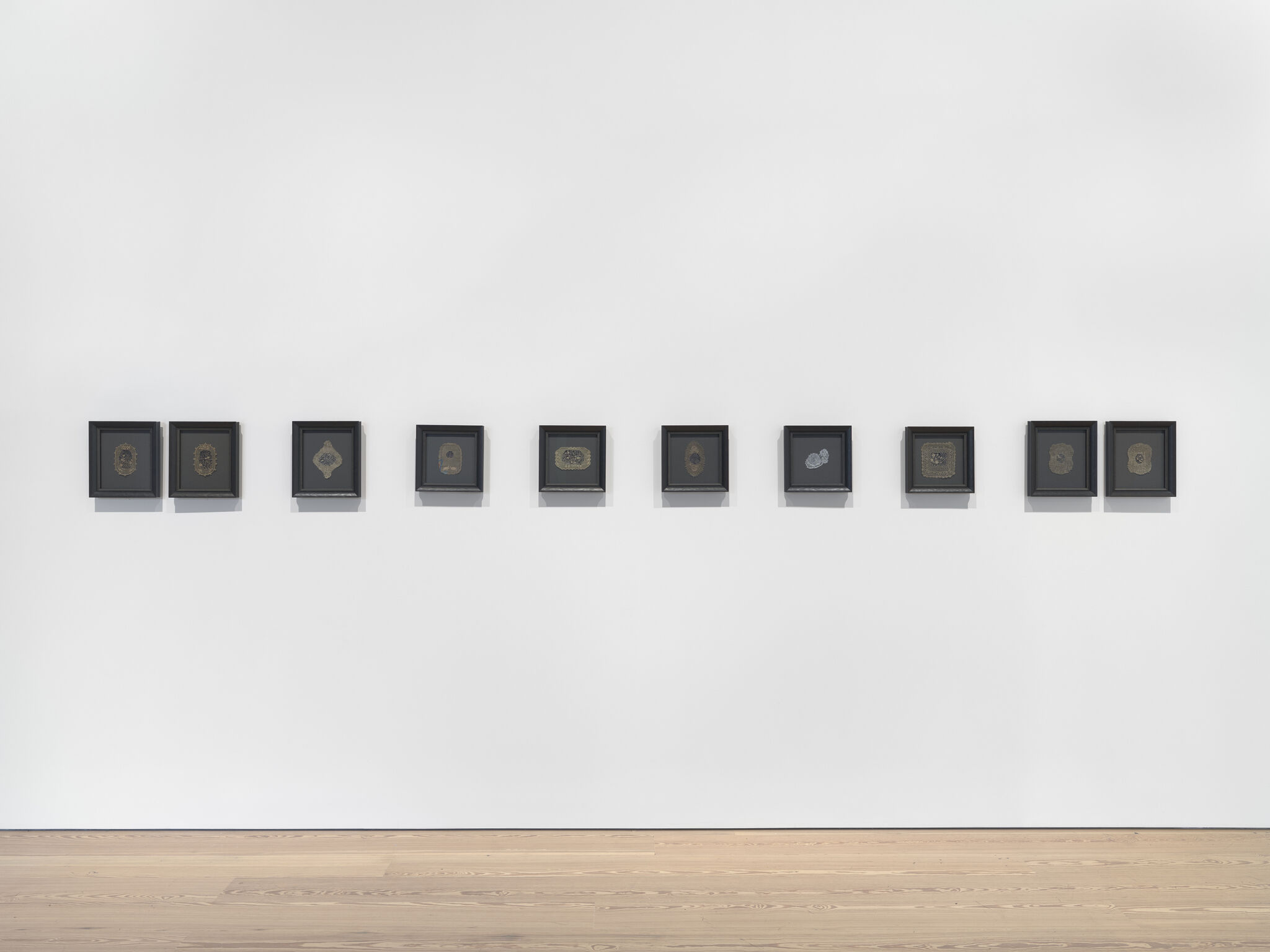 Small, intricate doilies in various black frames with grey backgrounds mounted onto a white wall.