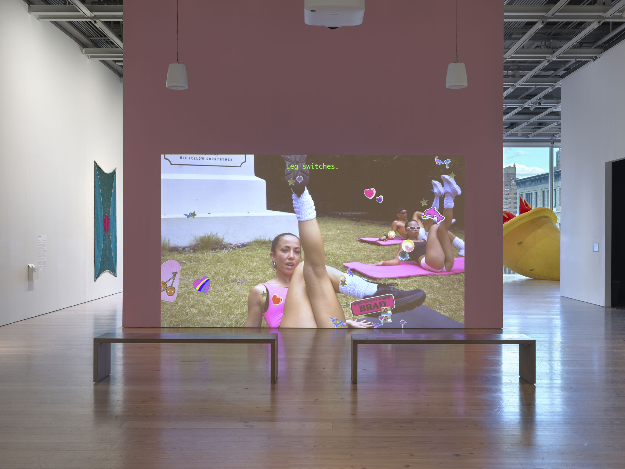 Two benches in front a video that is projected onto a blank wall. Women wearing pink leg warmers and 80's era exercise clothing do exercises with emoticons floating over the screen.