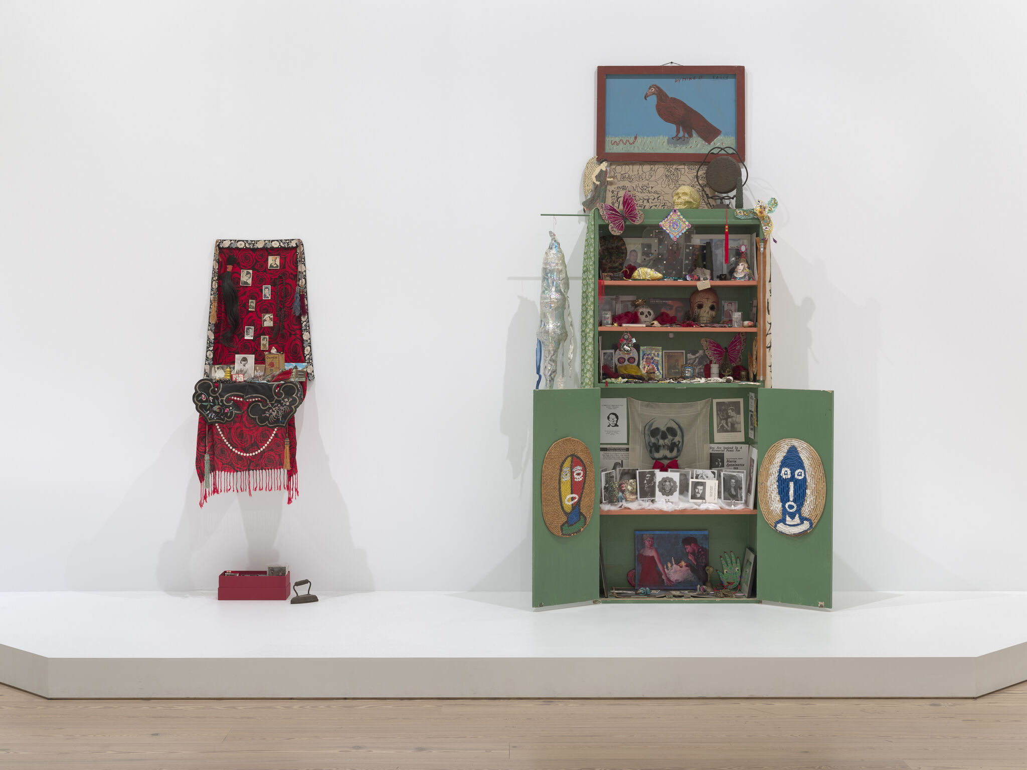 Two altars against a white wall. On the left, a smaller red altar with black and white photos and red fabric with fringe. On the right, a larger green altar with a painting with a brown bird against a blue sky and cabinet details with various items inside and the doors open.