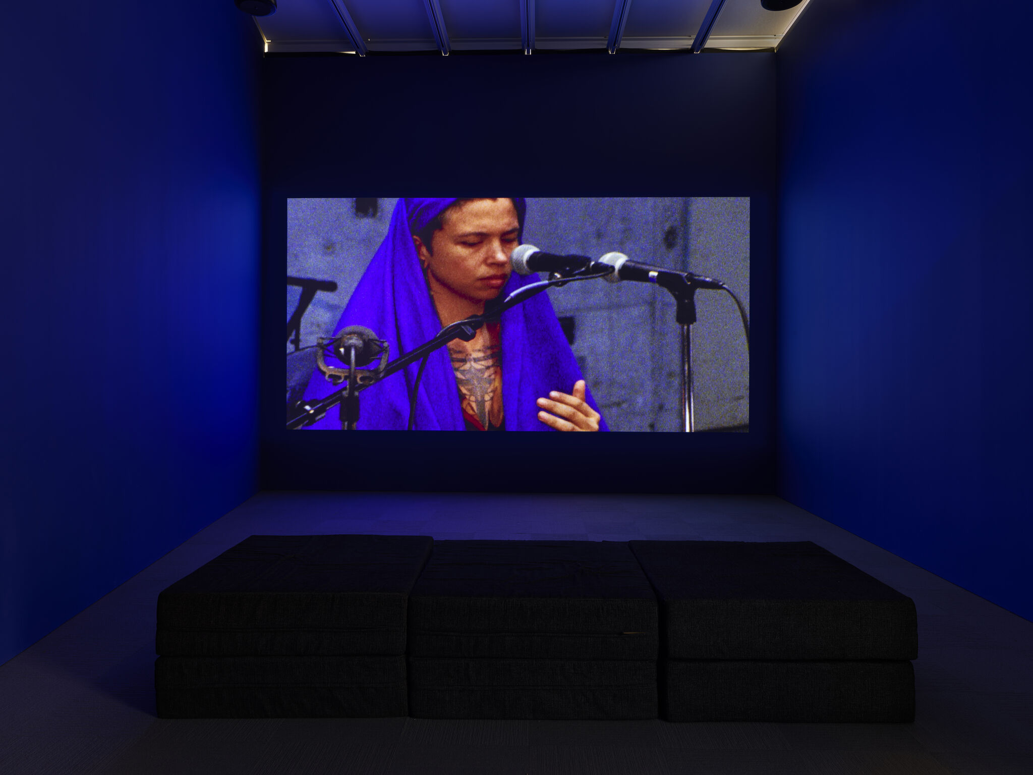 A long upholstered bench in a dark room with a projection on the wall. The projected image is a woman wrapped in purple talking into a microphone.