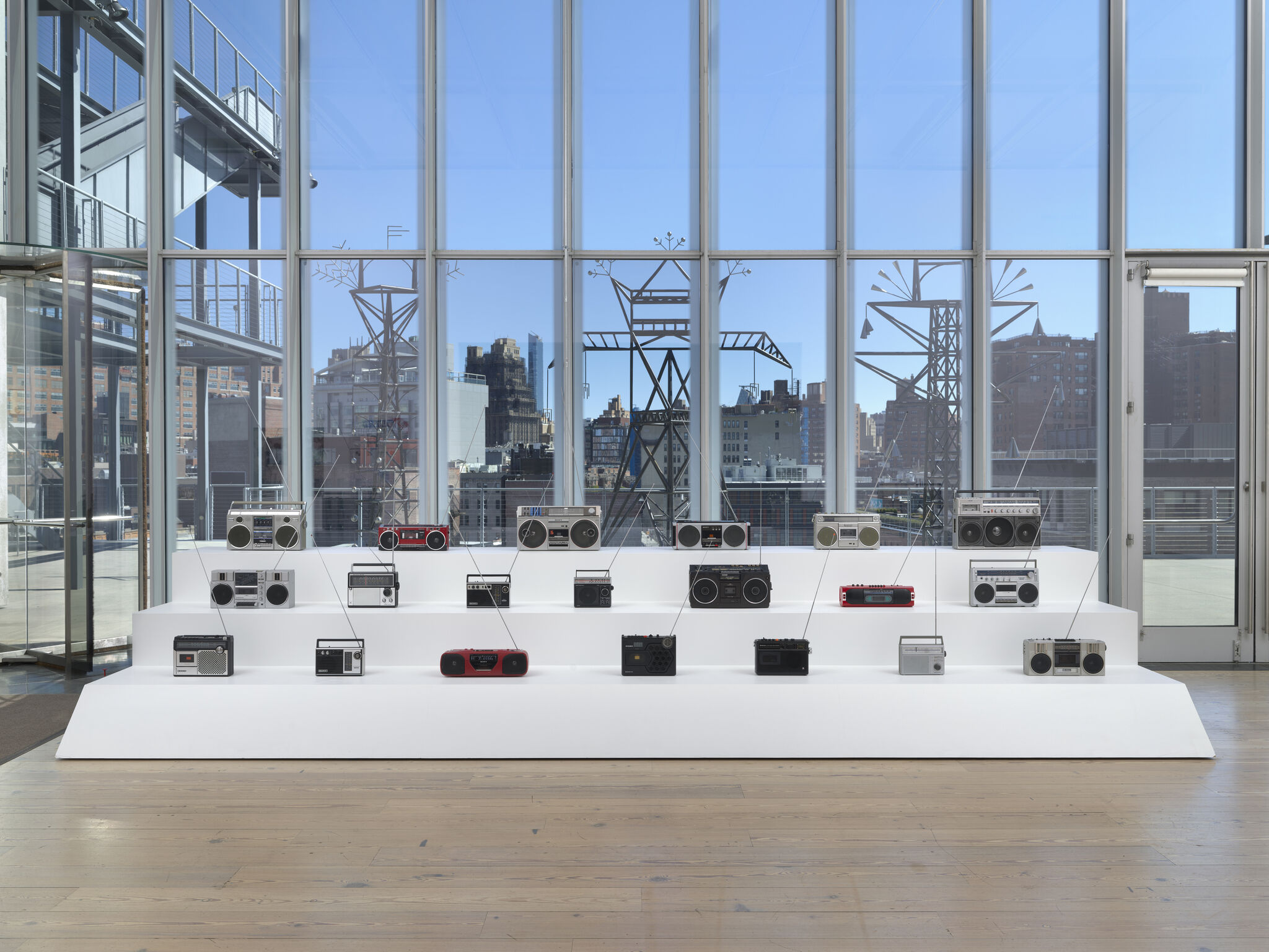 Rows of vintage radios and boomboxes displayed on white platforms in a glass-walled gallery.