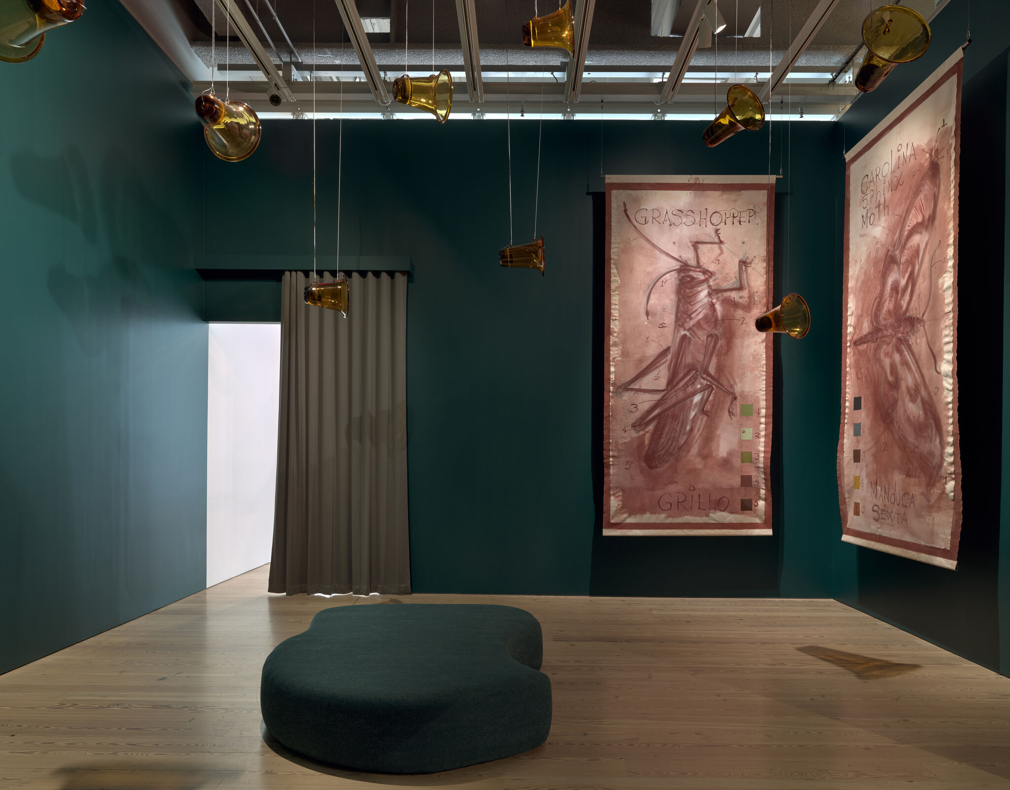 A room with curtains separating it from other rooms and deep turquoise walls. Glass-blown trumpet-shaped speakers hang from the ceiling by string and on the wall are canvas paintings with a dark red paint of grasshoppers.