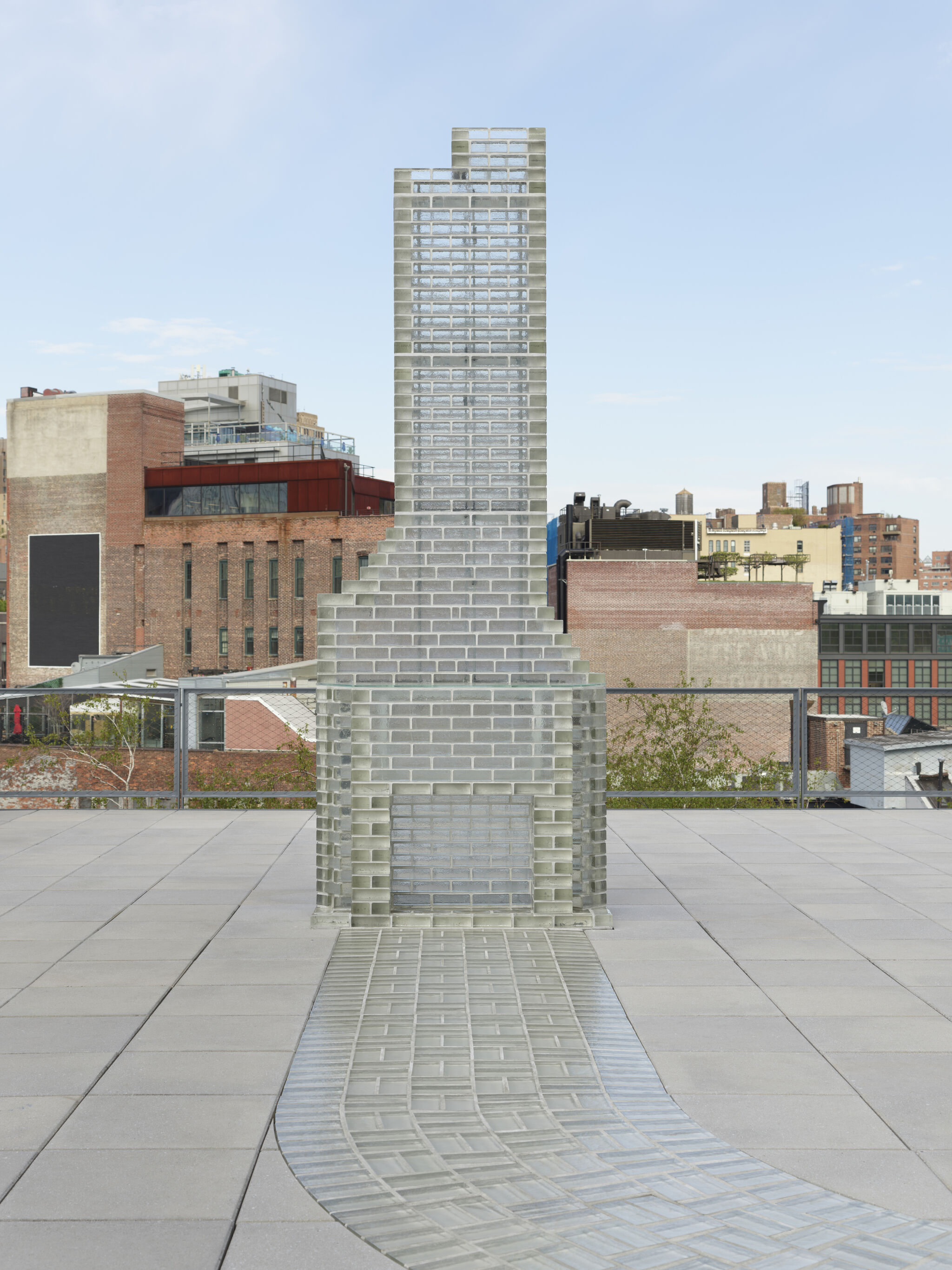 A tall glass-brick chimney sculpture stands on a rooftop with a reflective tile path leading to it.